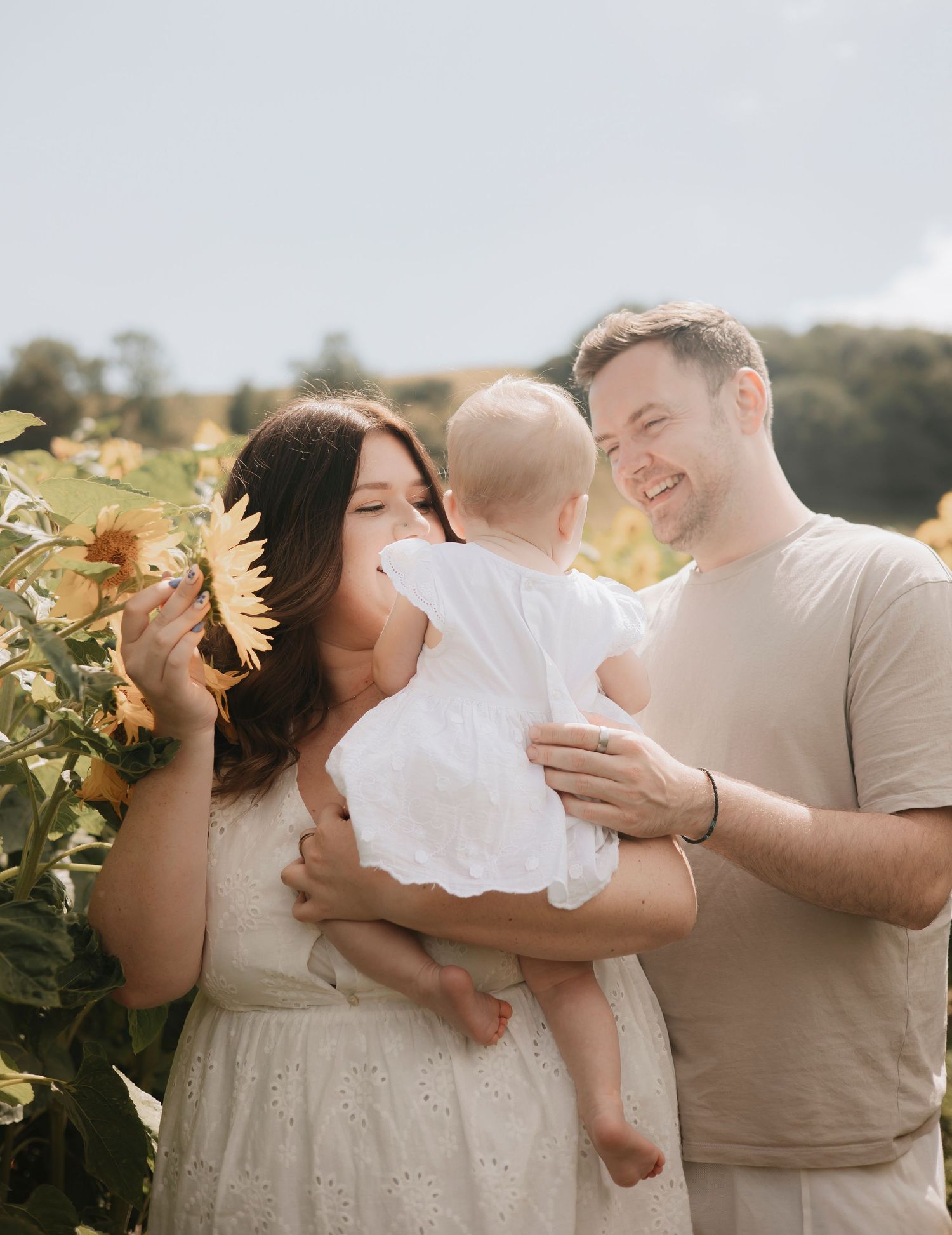 Happy family with mother, father, and baby in white dress posing in a sunny sunflower field.