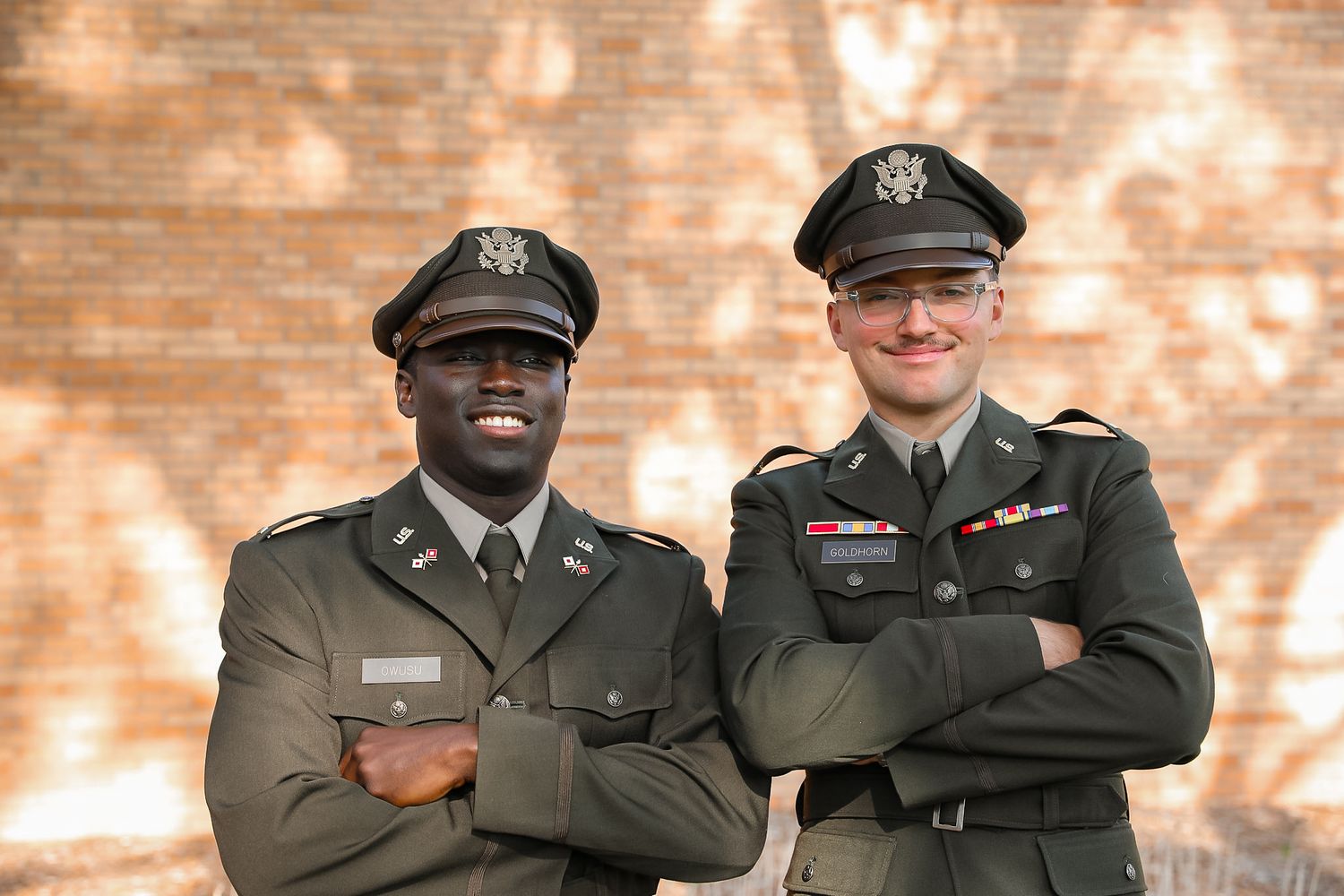 Two smiling military officers in green Army dress uniforms stand with arms crossed in front of a brick wall.