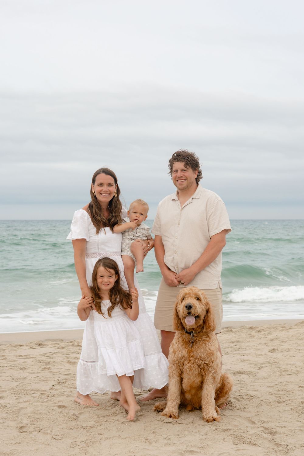 Family of four with a golden doodle dog posing on a sandy beach, smiling in white and beige outfits.