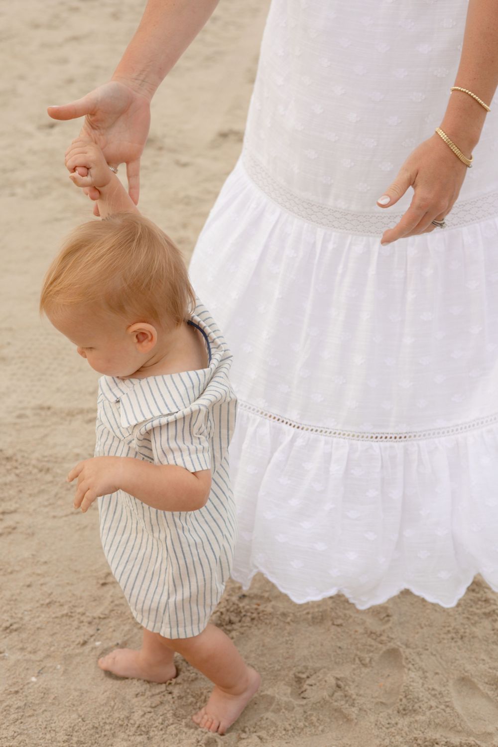 A mother in a white dress helps her baby take first steps on sandy beach, baby wearing striped romper.