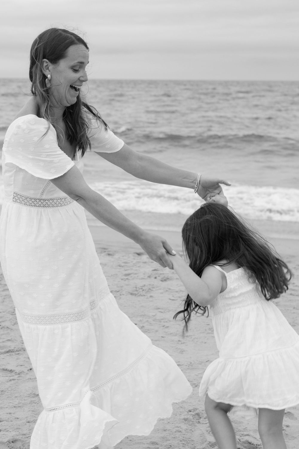 Mother and daughter in matching white dresses dancing joyfully on the beach in black and white photo.