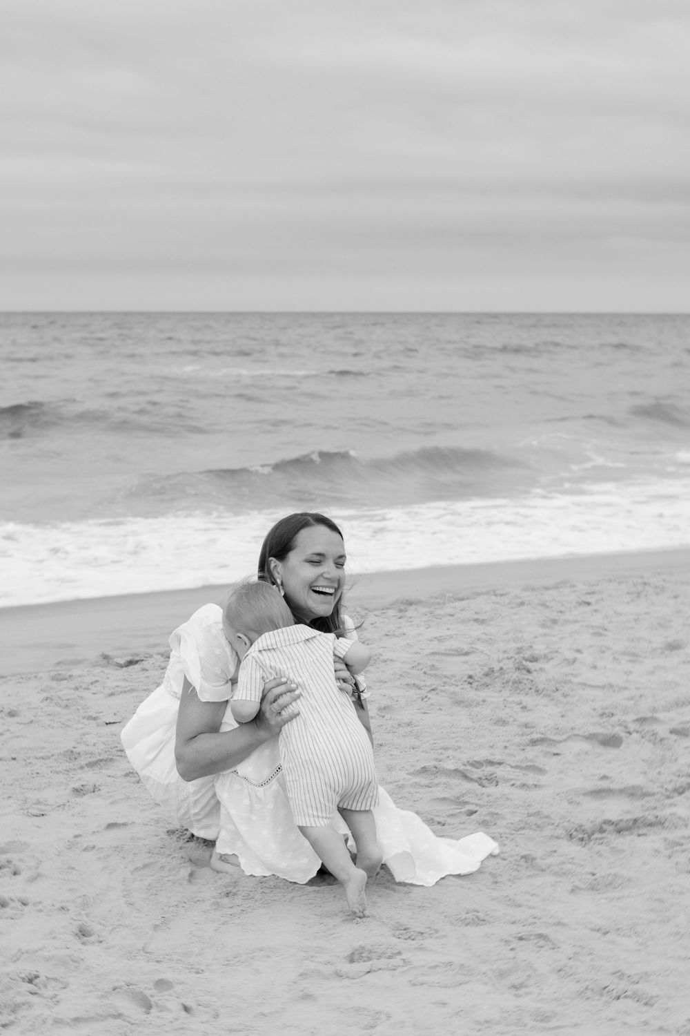Mother laughing while hugging baby on sandy beach in elegant white dress, black and white photo.