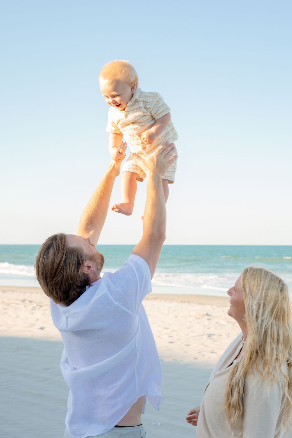 Father lifts smiling baby in the air on a sunny beach while blonde mother watches joyfully.
