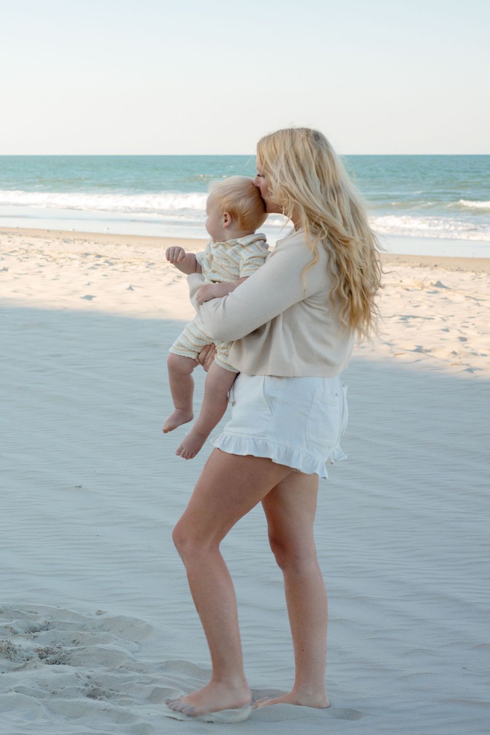 Blonde mother holding baby on sandy beach with ocean waves, wearing white skirt and beige top, barefoot.