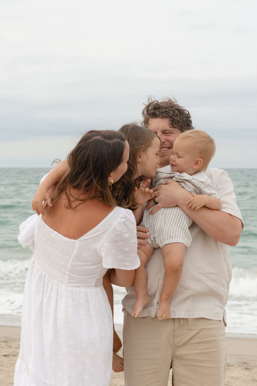 Family of four sharing a joyful moment on a sandy beach, parents embracing children with ocean waves in the background.