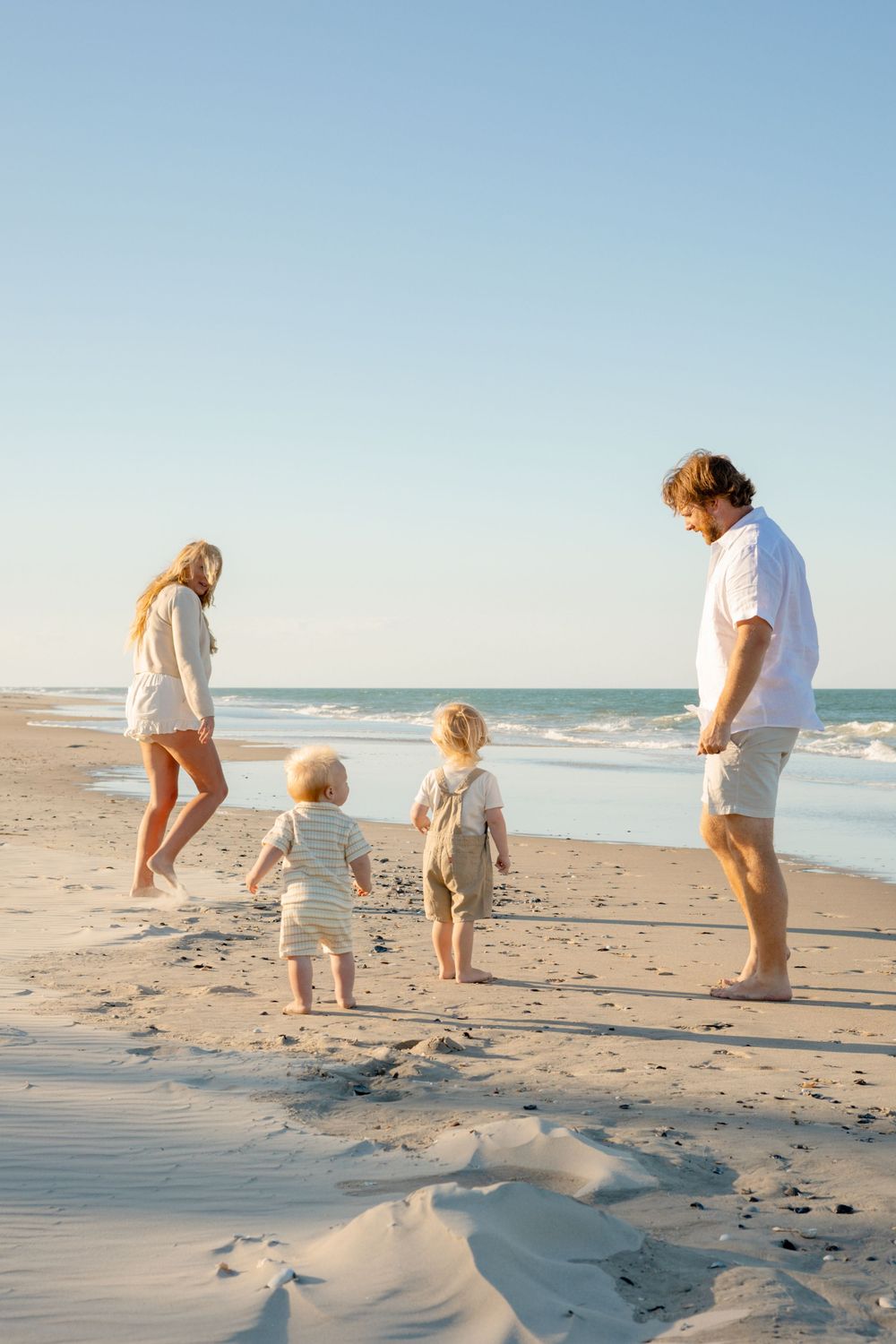 Family with two toddlers enjoying a sunny beach walk on white sand with ocean waves in the background.