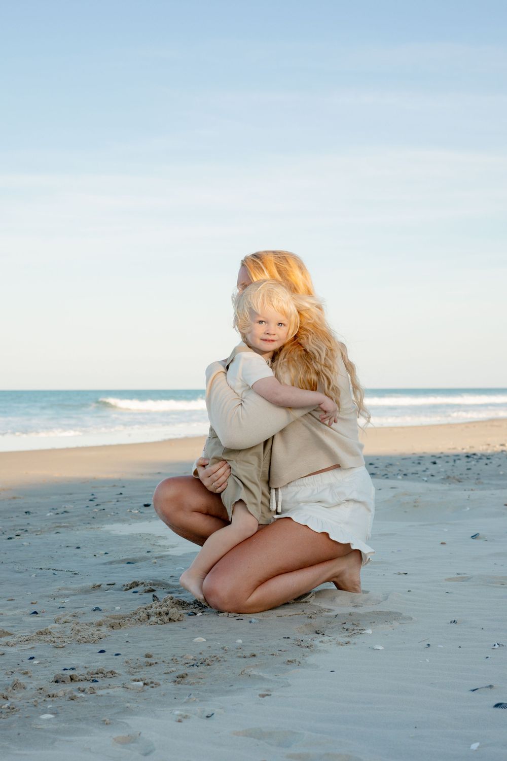 A mother with long blonde hair kneels on a sandy beach, hugging her young blonde toddler by the ocean.