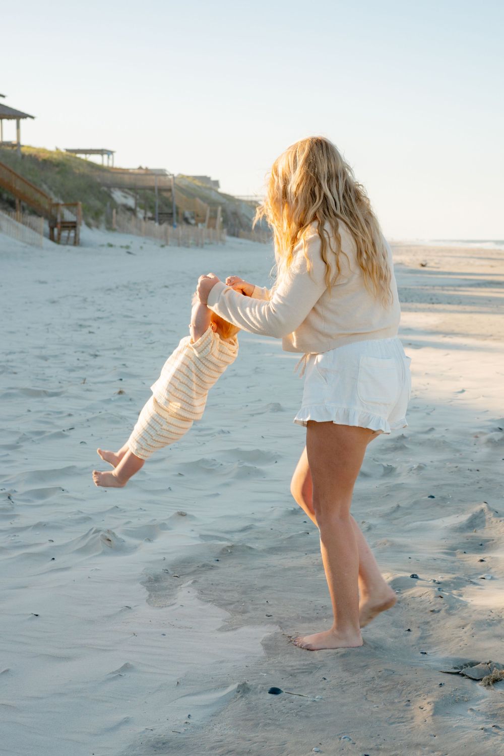 A blonde woman playfully lifts a baby on a sunny sandy beach, both barefoot near coastal dunes.