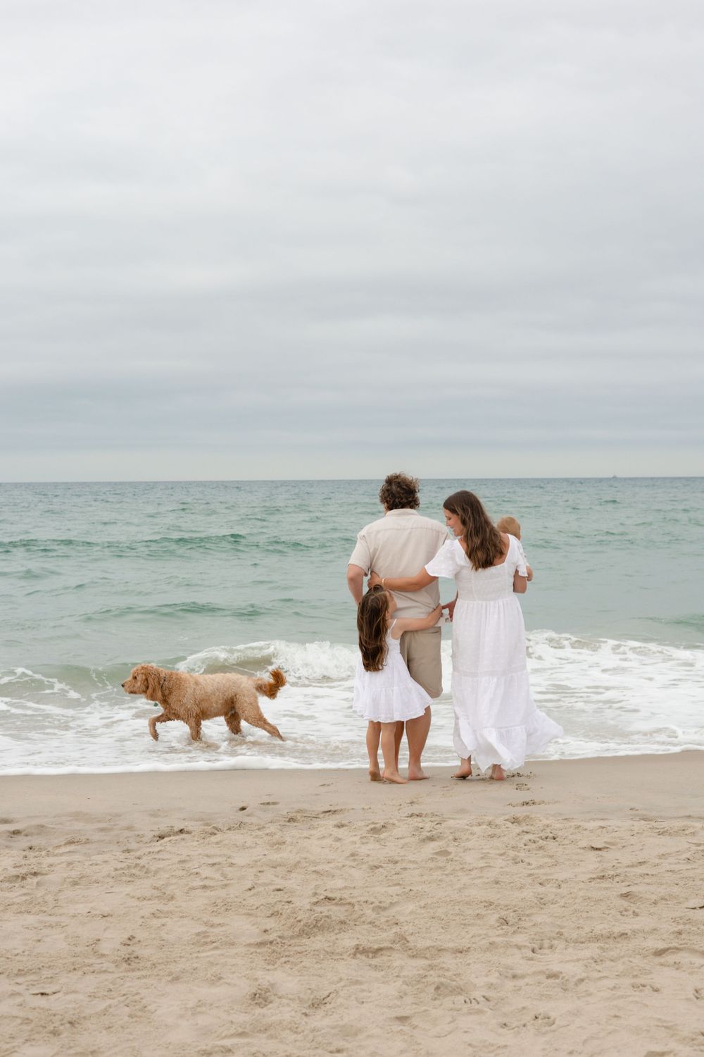 Family with dog enjoying a walk on the beach, with waves and overcast skies in the background.