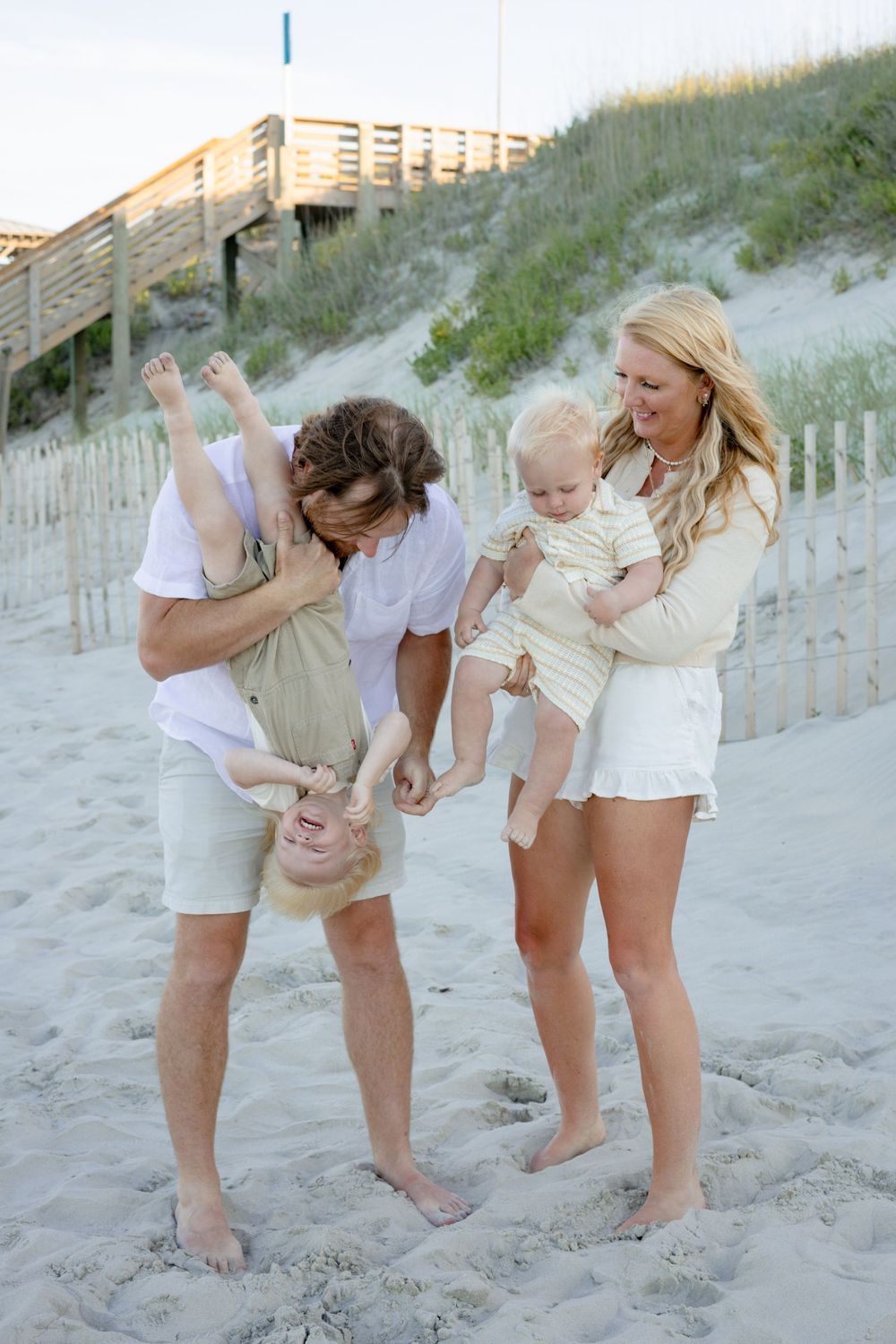 Happy family of four enjoying a playful moment on a sandy beach, with dad holding child upside down.