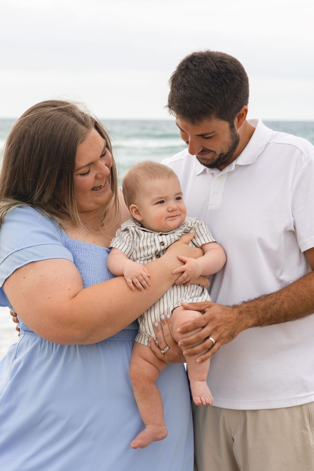 Happy young family with baby at the beach, parents smiling at infant in blue dress and white polo.