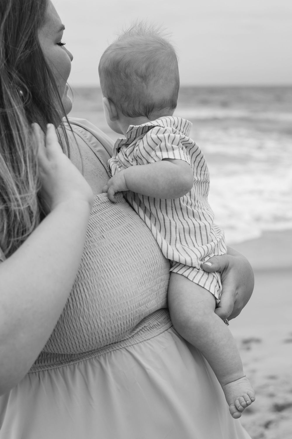 Black and white photo of a mother holding her baby on a beach, with ocean waves in the background.
