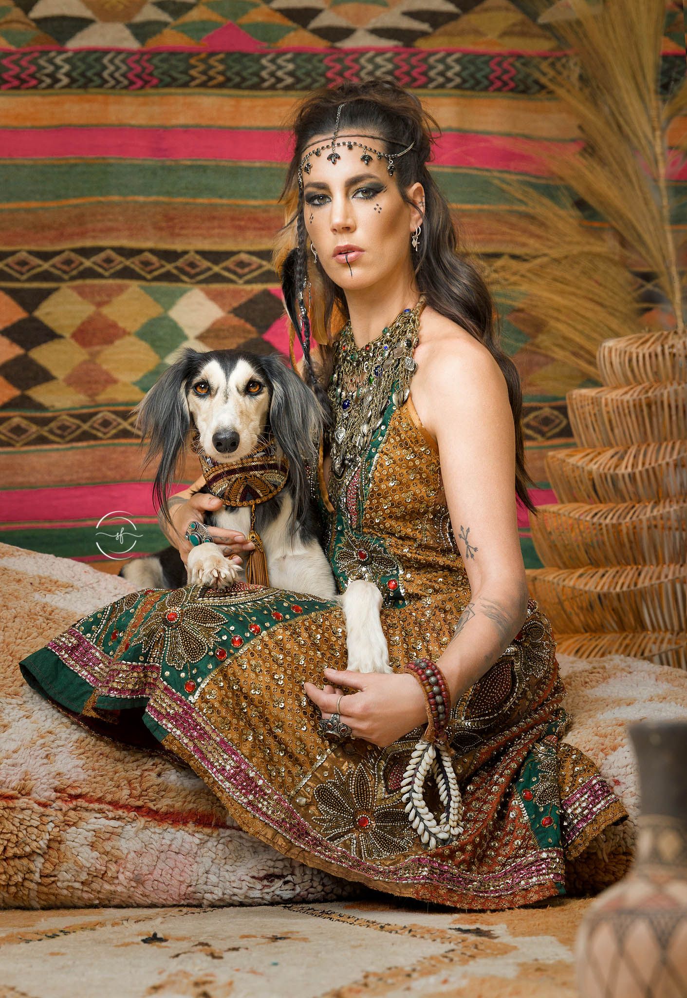 Woman in ornate golden ethnic dress with dramatic makeup poses with a Saluki dog on a patterned rug.