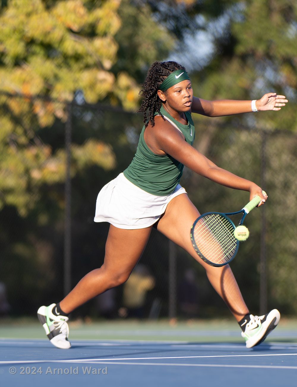 Tennis player in green tank top and white skirt hits forehand shot on outdoor court during competition.