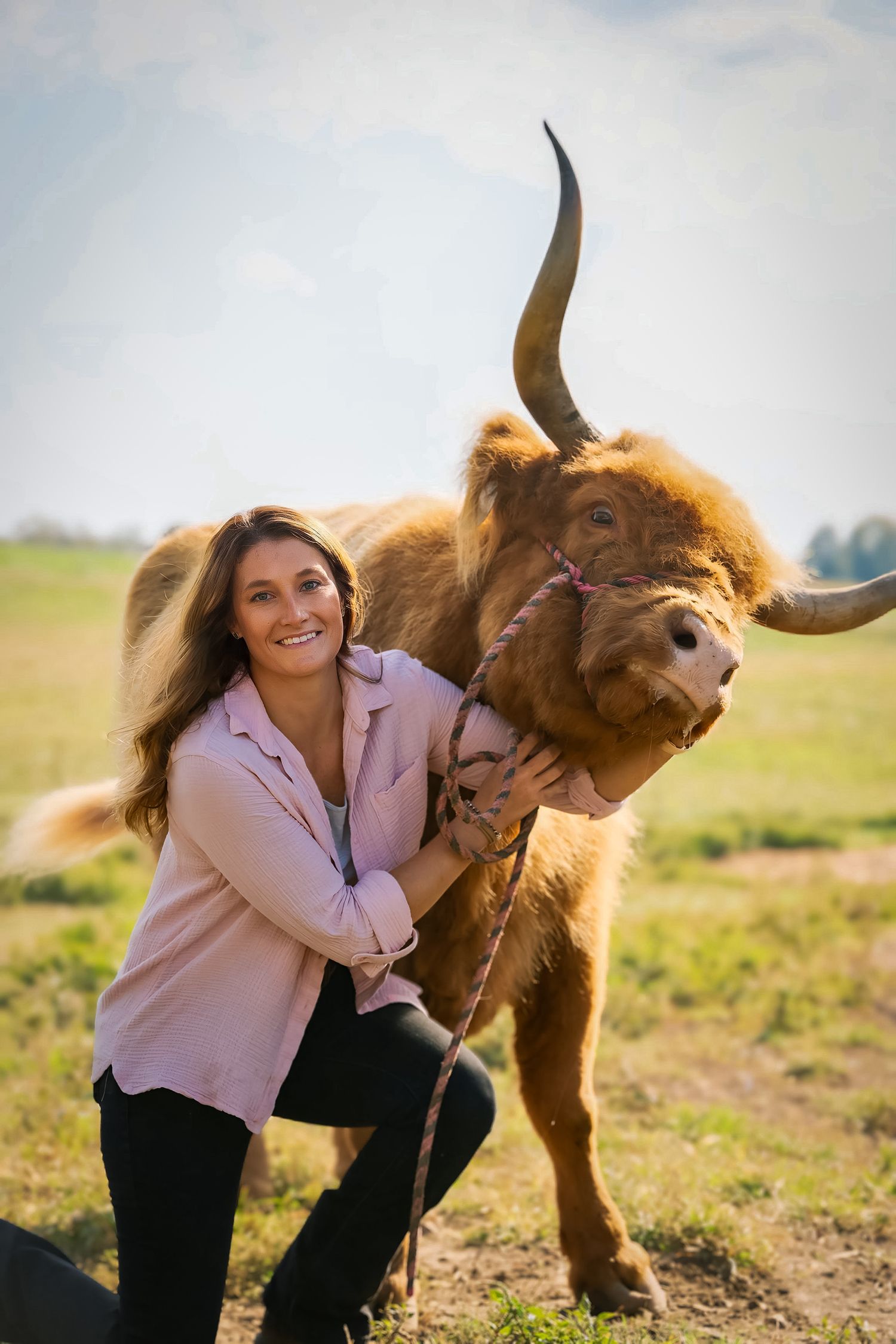 Smiling woman hugging a fluffy Highland cow with long horns in a green field.