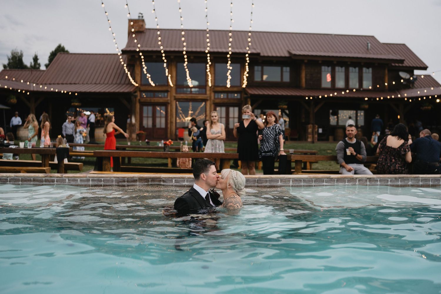Bride and groom share a romantic kiss in a pool at their outdoor wedding reception with string lights.