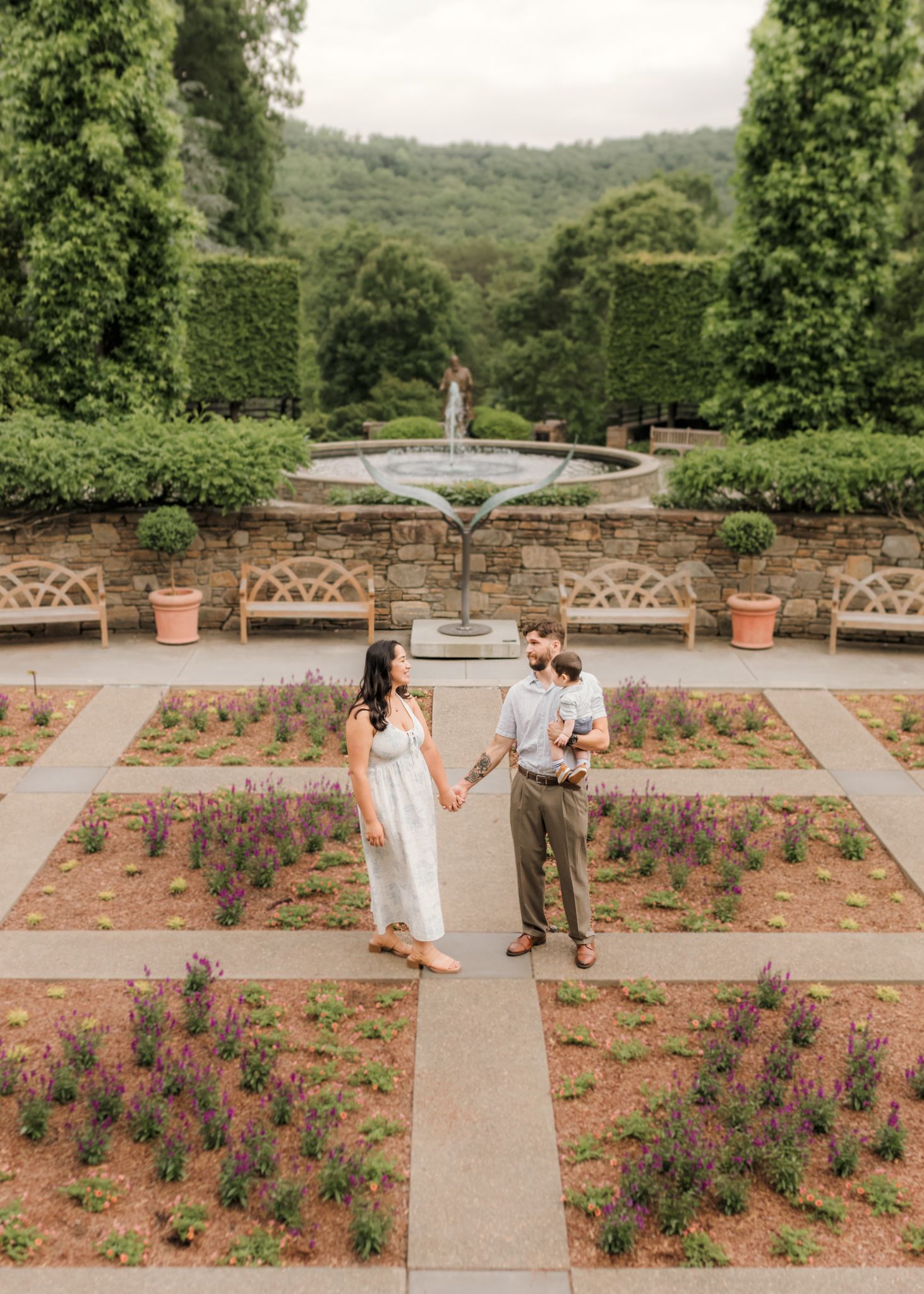 Couple holds hands in a formal garden with a fountain, lush hedges, and colorful flower beds on a cloudy day.
