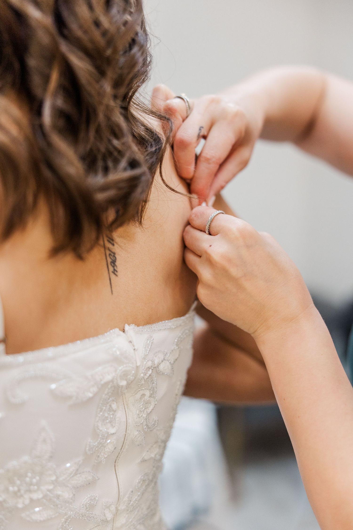 Bridesmaid fastening the back of a lace wedding dress on a bride with a tattoo on her shoulder blade.