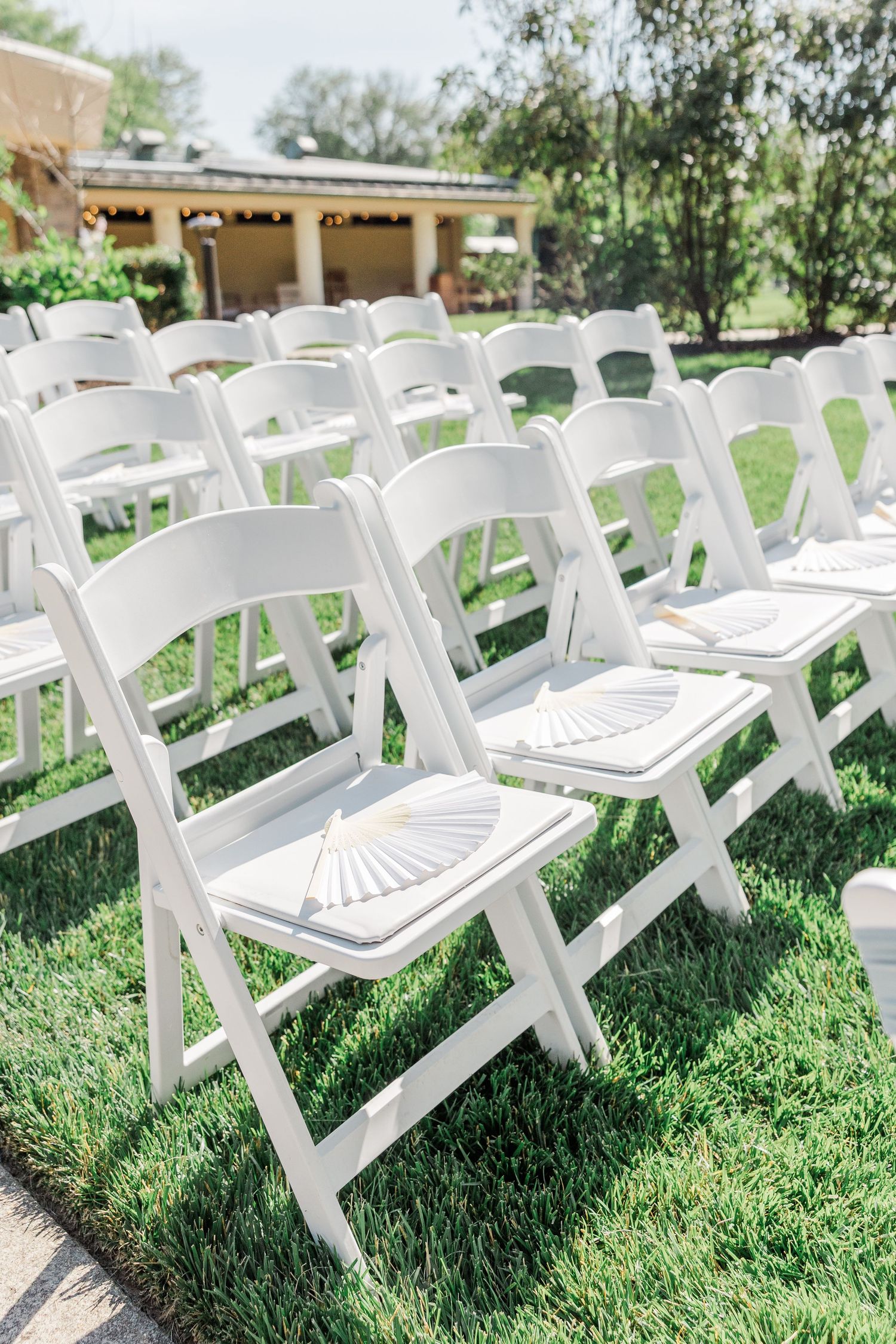 Rows of white folding chairs with paper fans on green grass for an outdoor wedding ceremony.