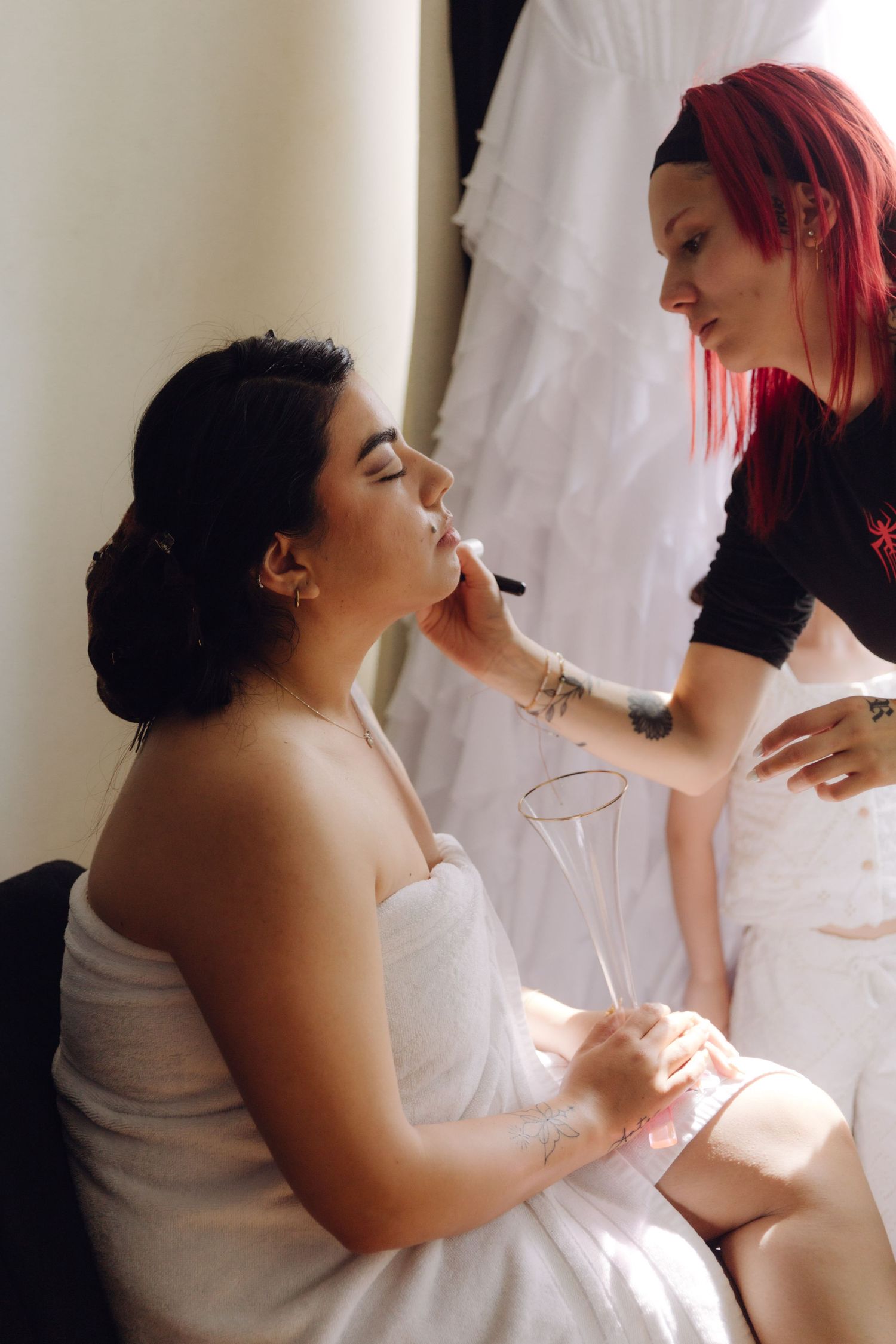 Professional makeup artist with red hair applies makeup to bride holding a champagne flute.
