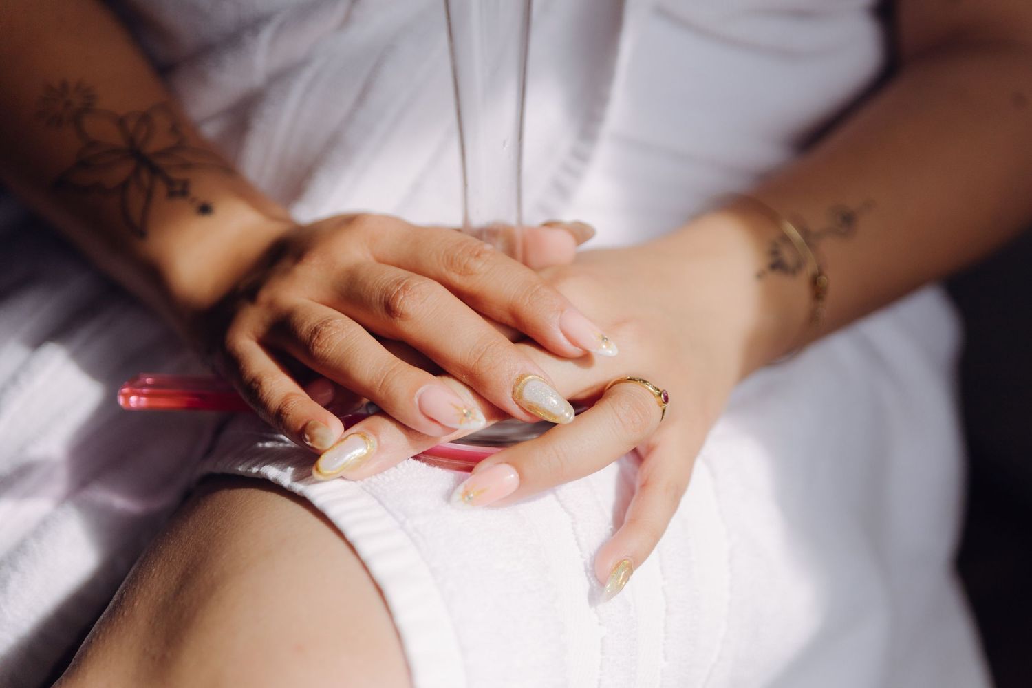 Close-up of bride's hands with tattoos, gold ring, and pastel manicure resting on white fabric.