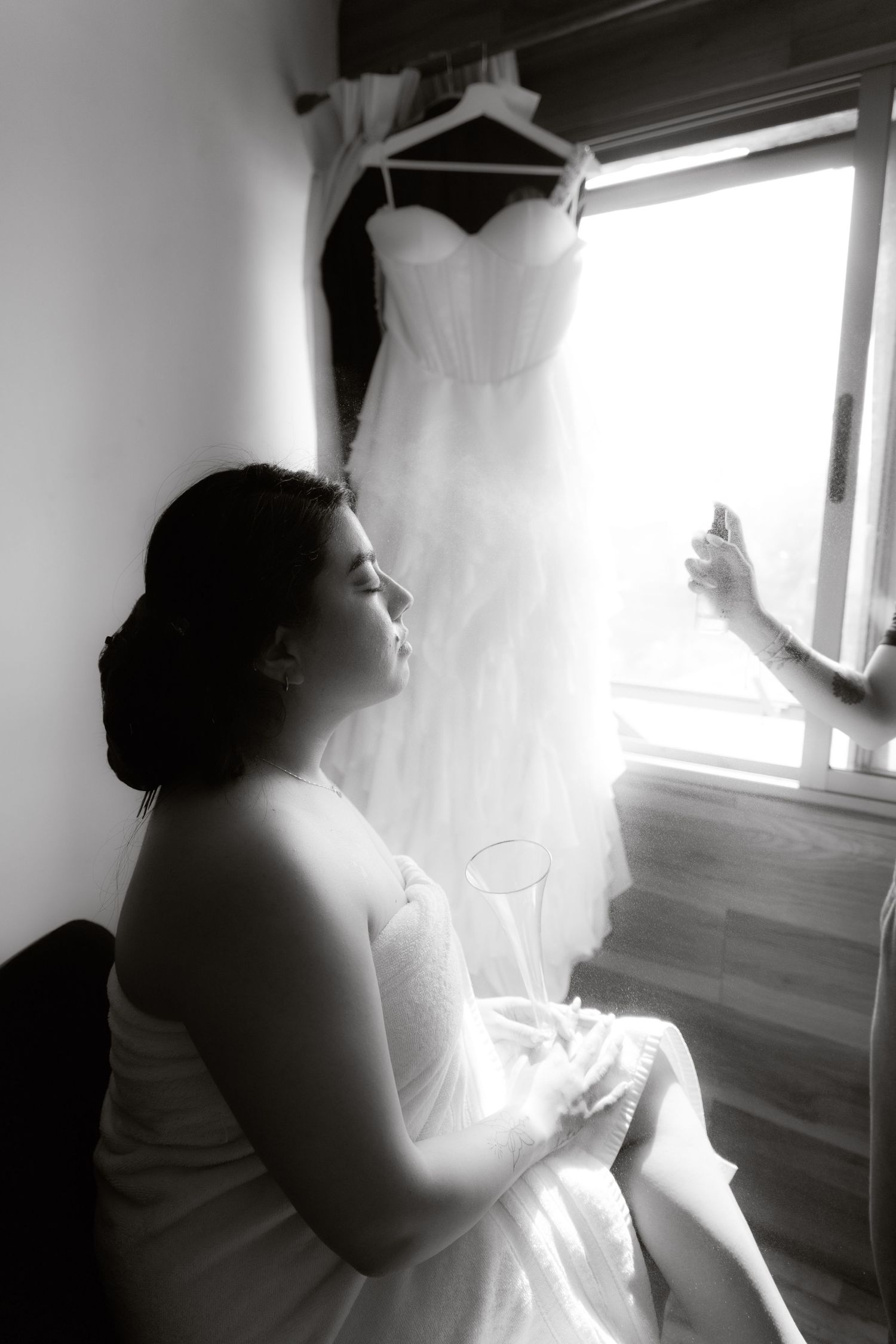 Black and white portrait of bride gazing at her wedding dress hanging by a sunlit window.
