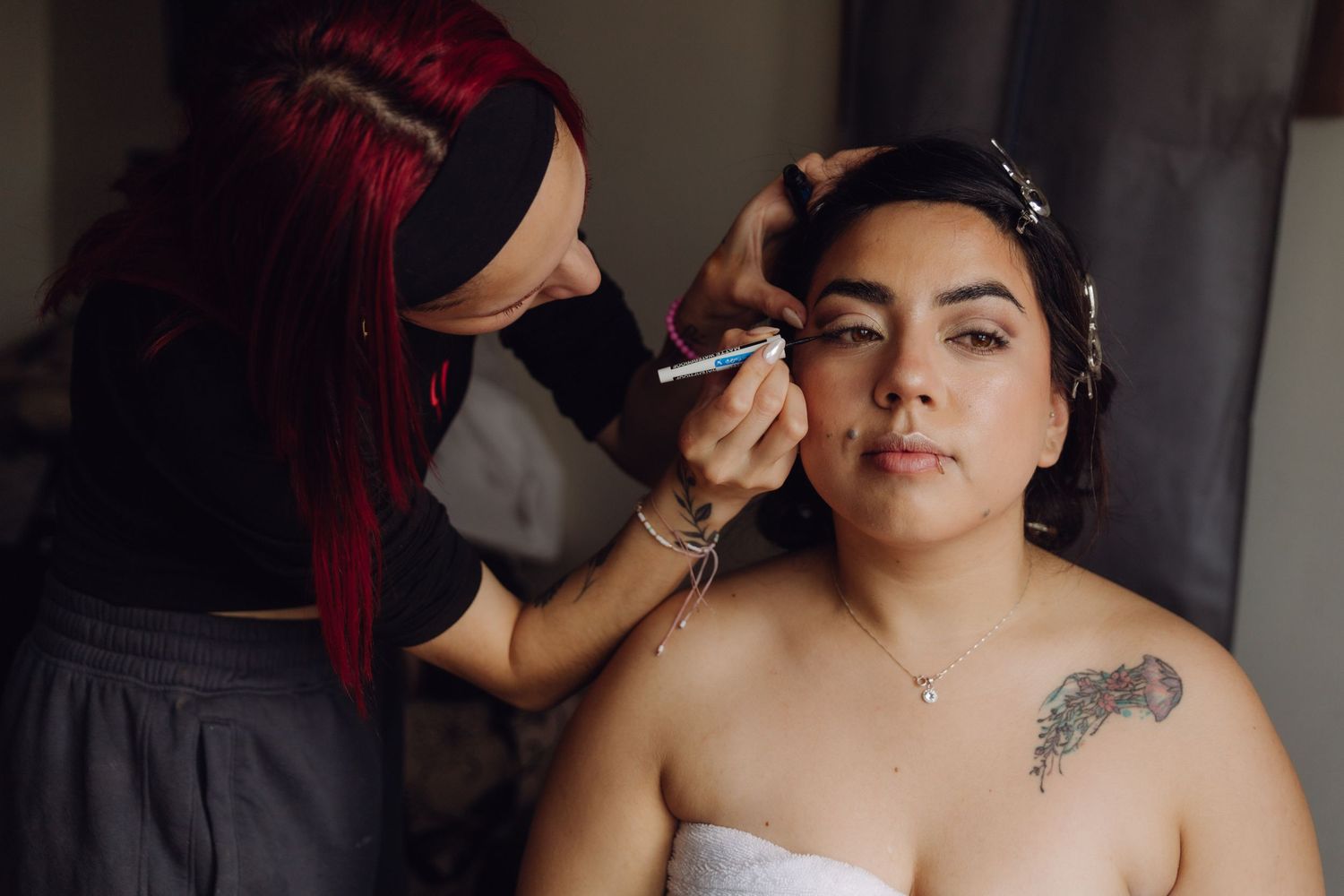 Makeup artist carefully applies eye makeup to tattooed bride wrapped in white towel.