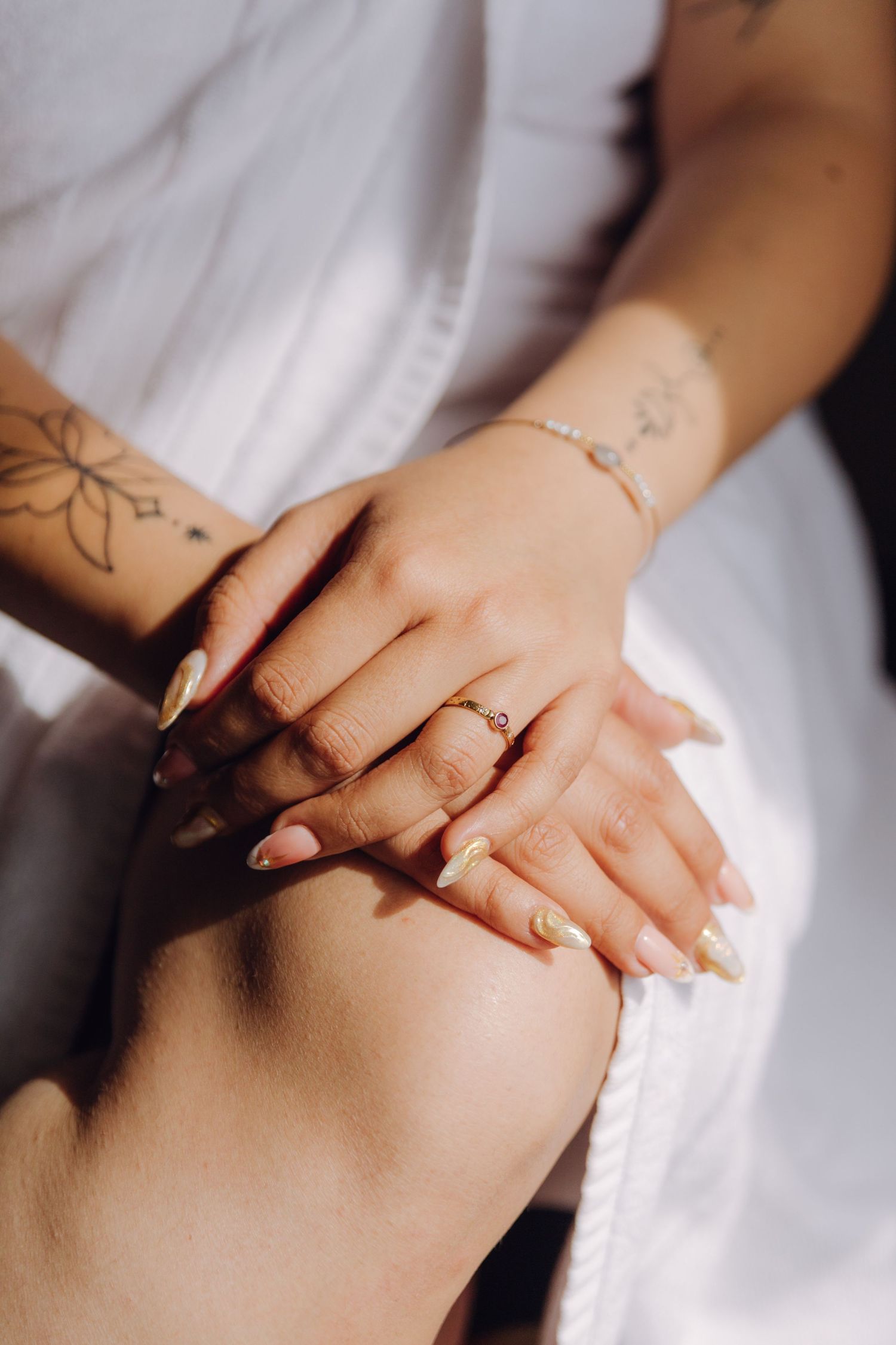 Close-up of bride's tattooed hands with gold ring and elegant manicure resting on white fabric.