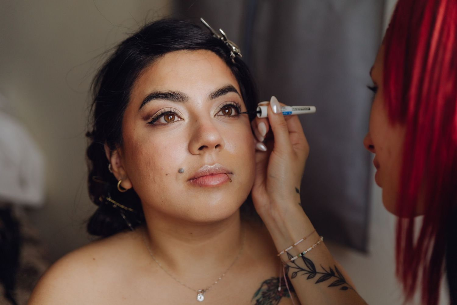 Professional makeup artist applying eye makeup to young brunette bride with tattoos and necklace.