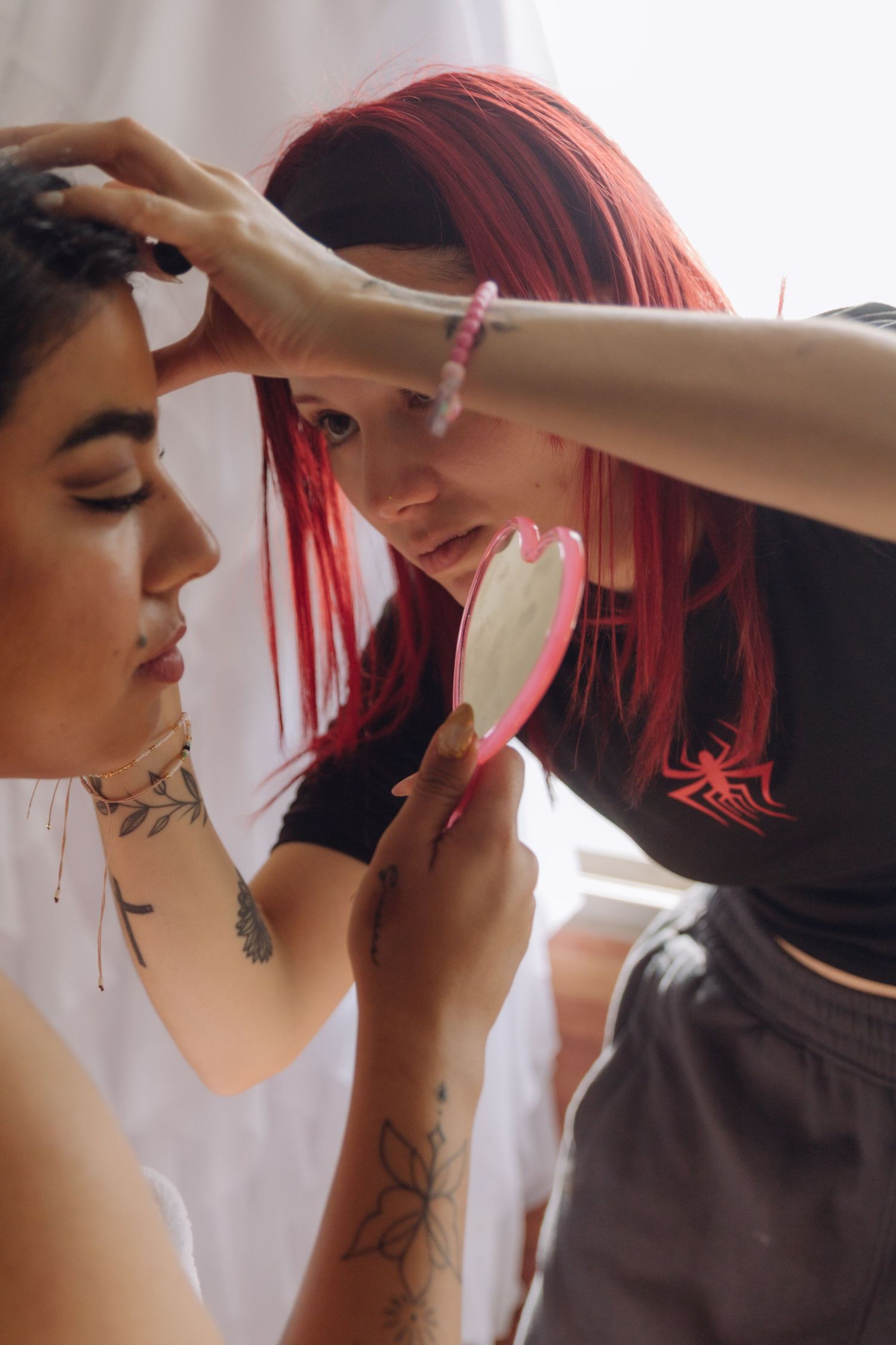 Red-haired makeup artist holding pink mirror while styling dark-haired tattooed woman's hair and makeup.