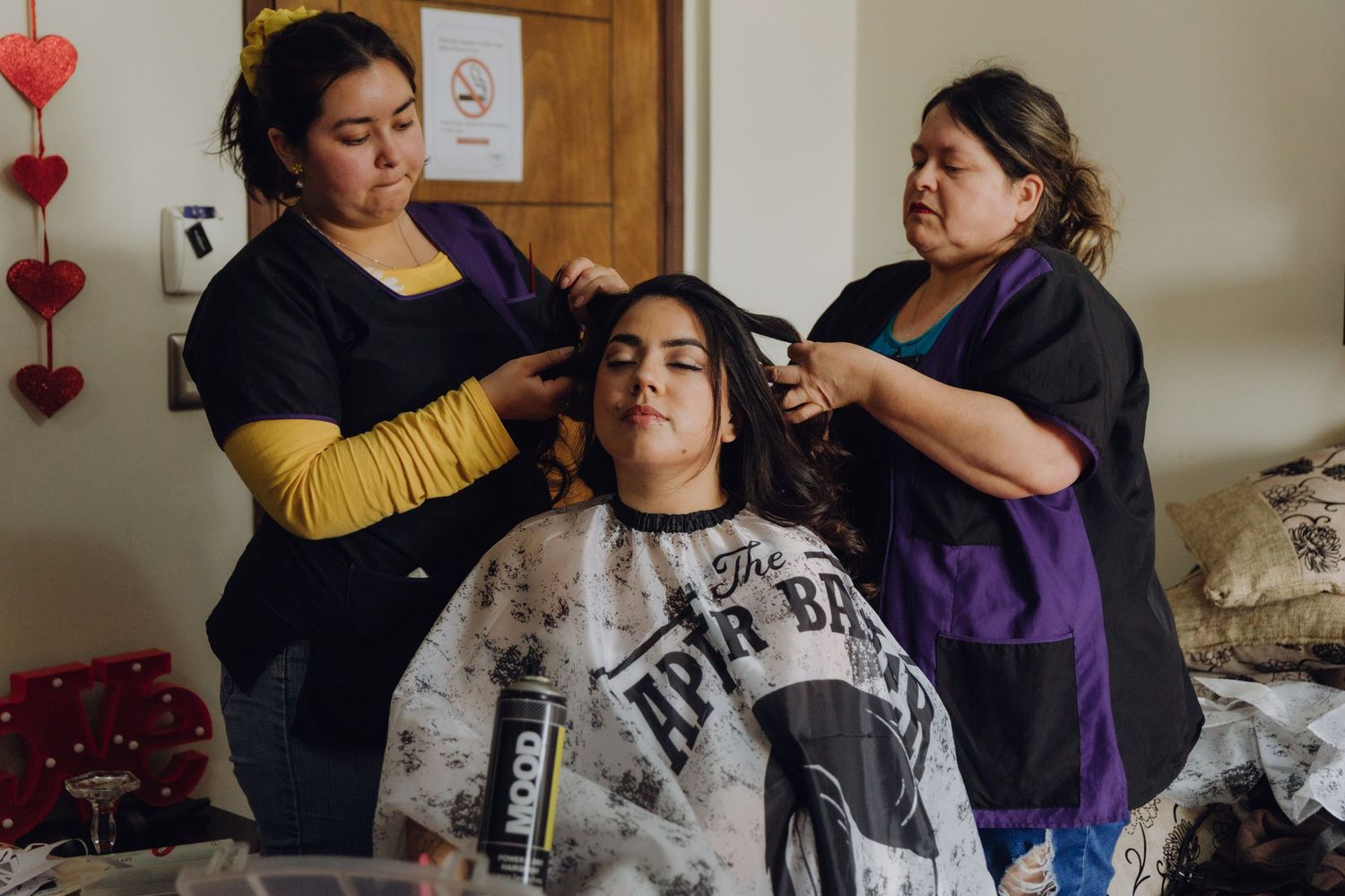 Two professional hairstylists working on bride's hair in salon cape during wedding preparation.