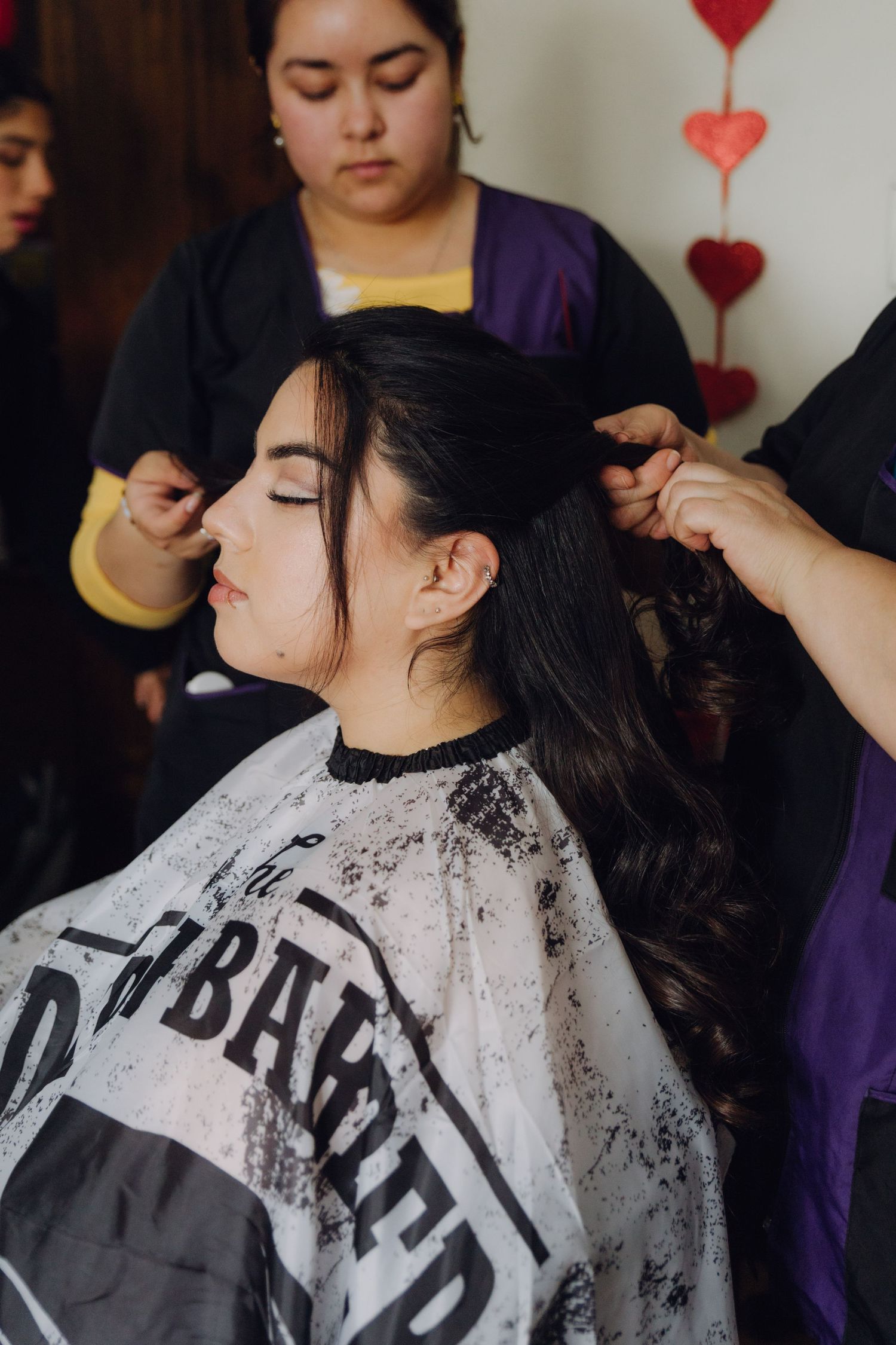 Two hairstylists in black and purple aprons styling long dark hair of seated woman in salon cape.