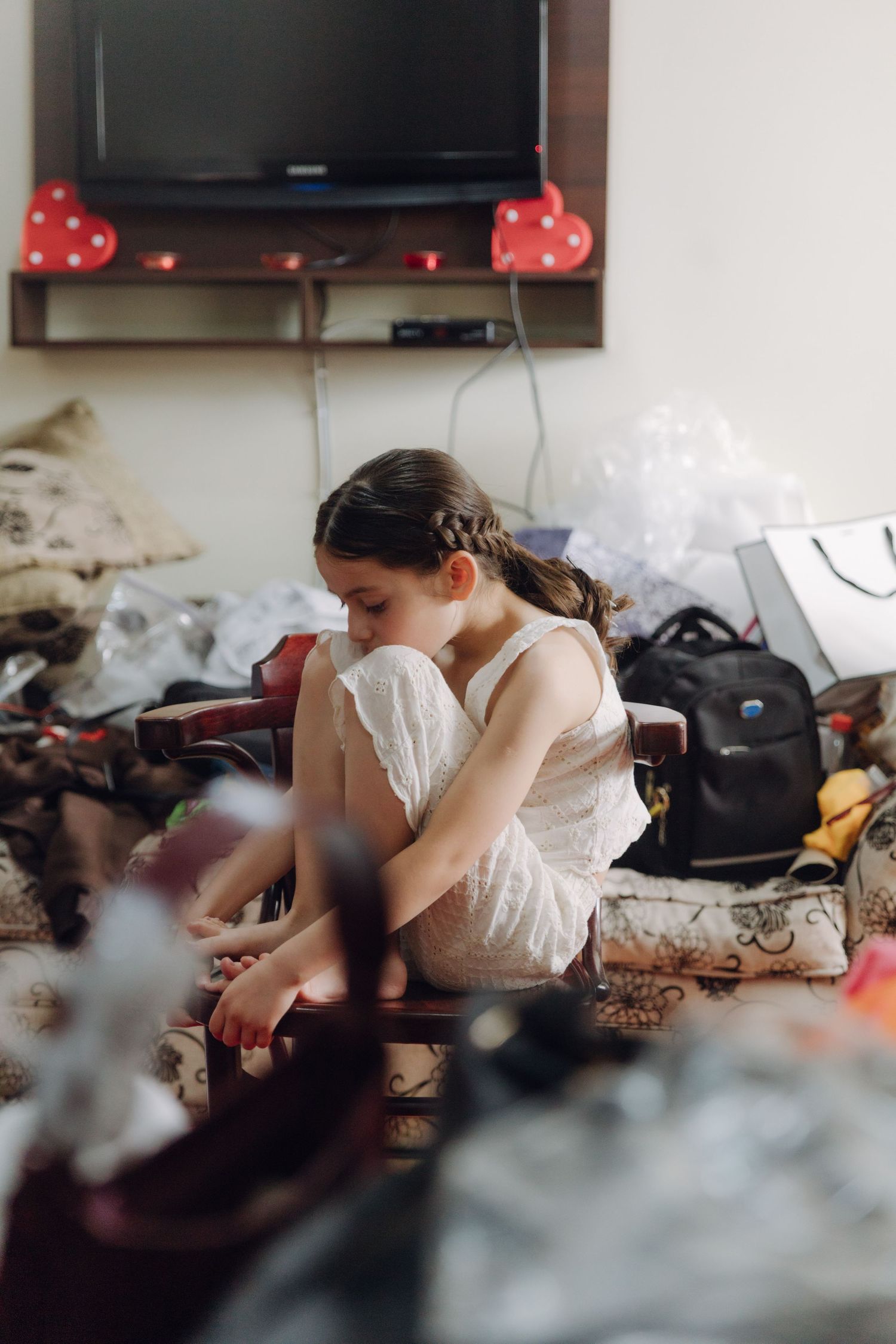 Young girl in white lace dress sitting on chair surrounded by wedding preparation items.