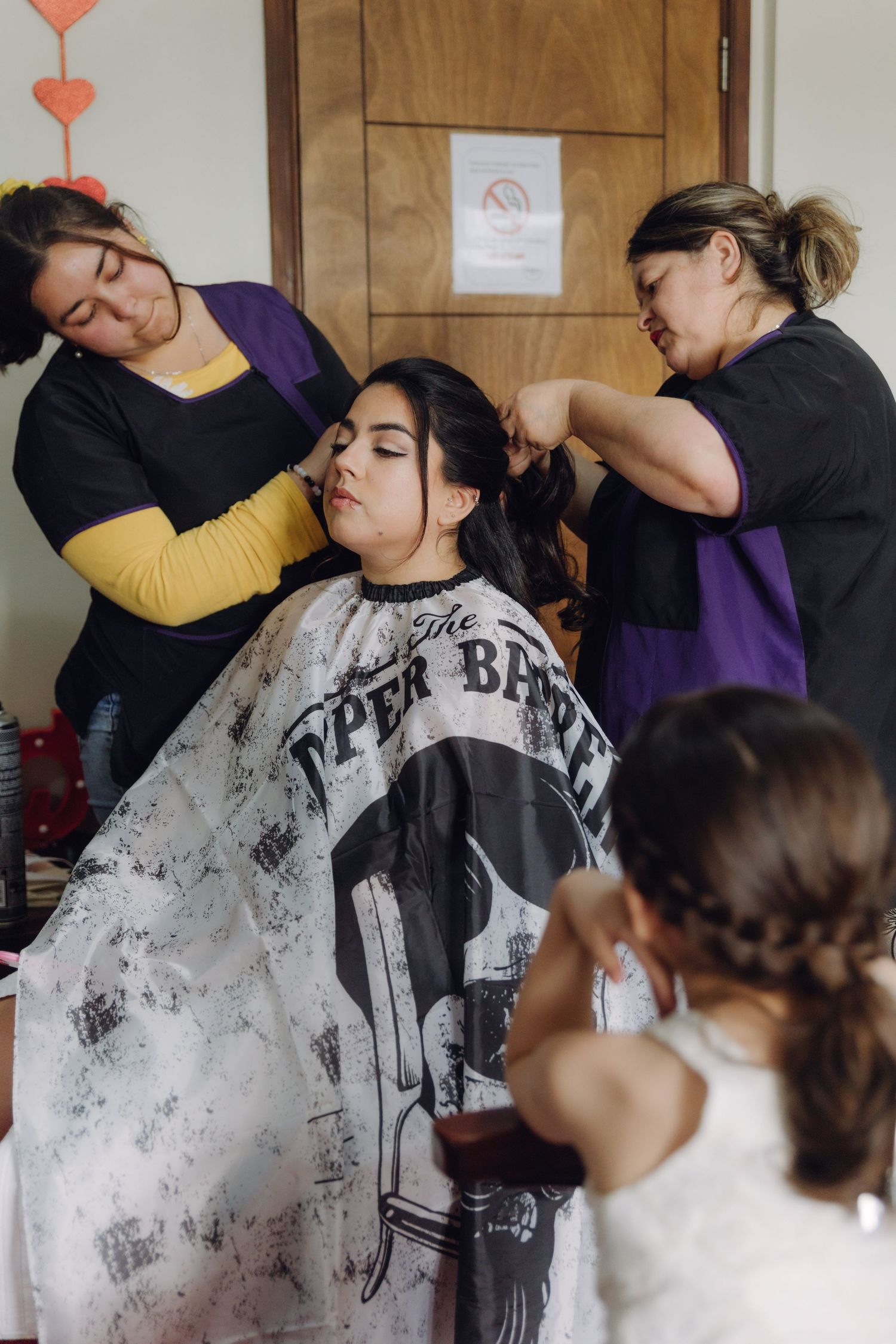 Two hairstylists in matching purple aprons styling seated bride's hair during wedding preparations.