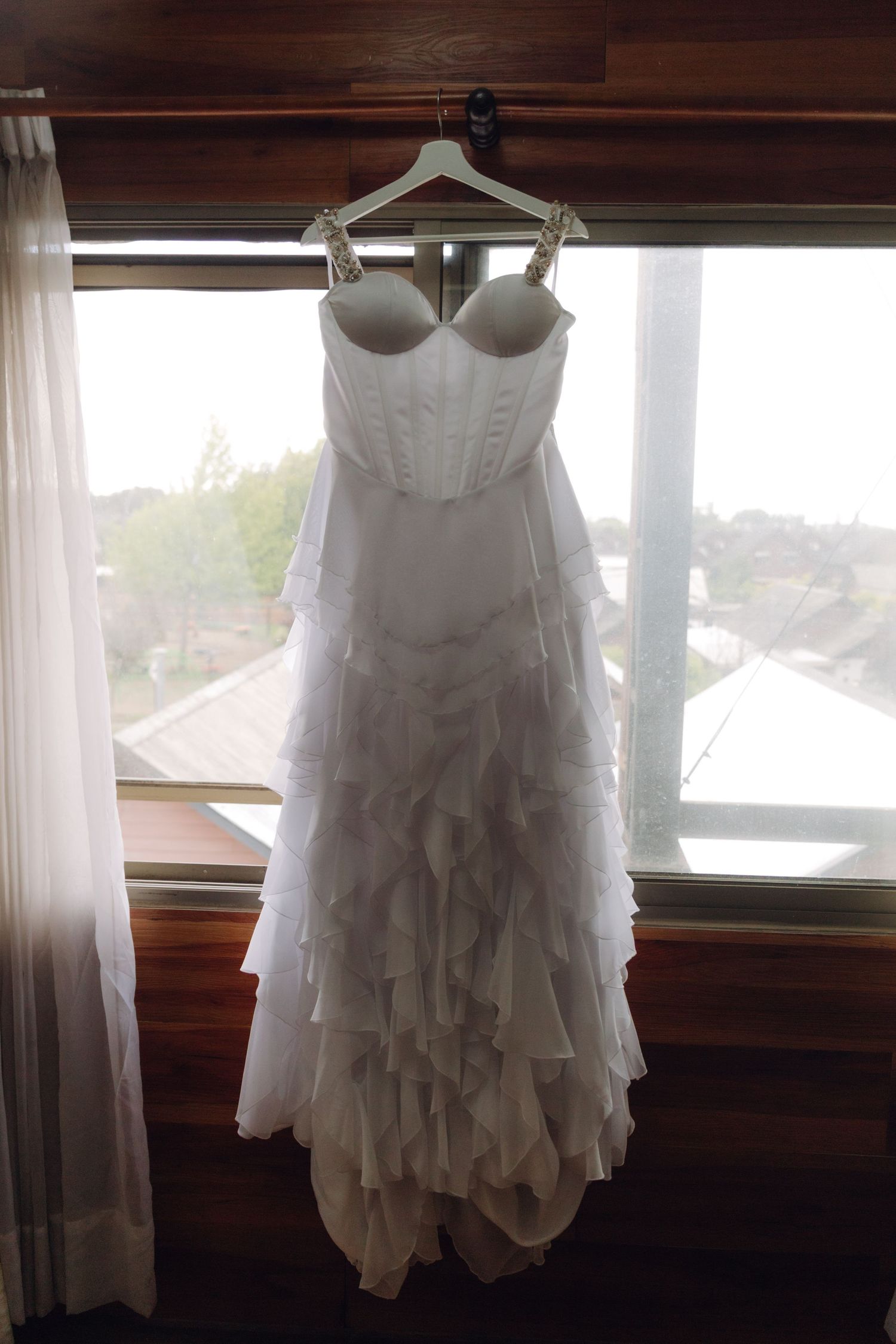Elegant white strapless ruffled wedding dress hanging on hanger in front of bright window.