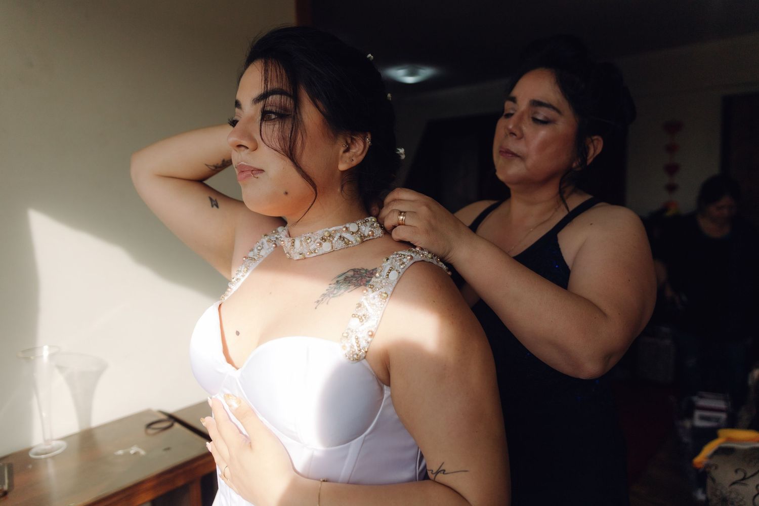 Woman helping bride fasten jeweled necklace while wearing white bridal bra during wedding prep.