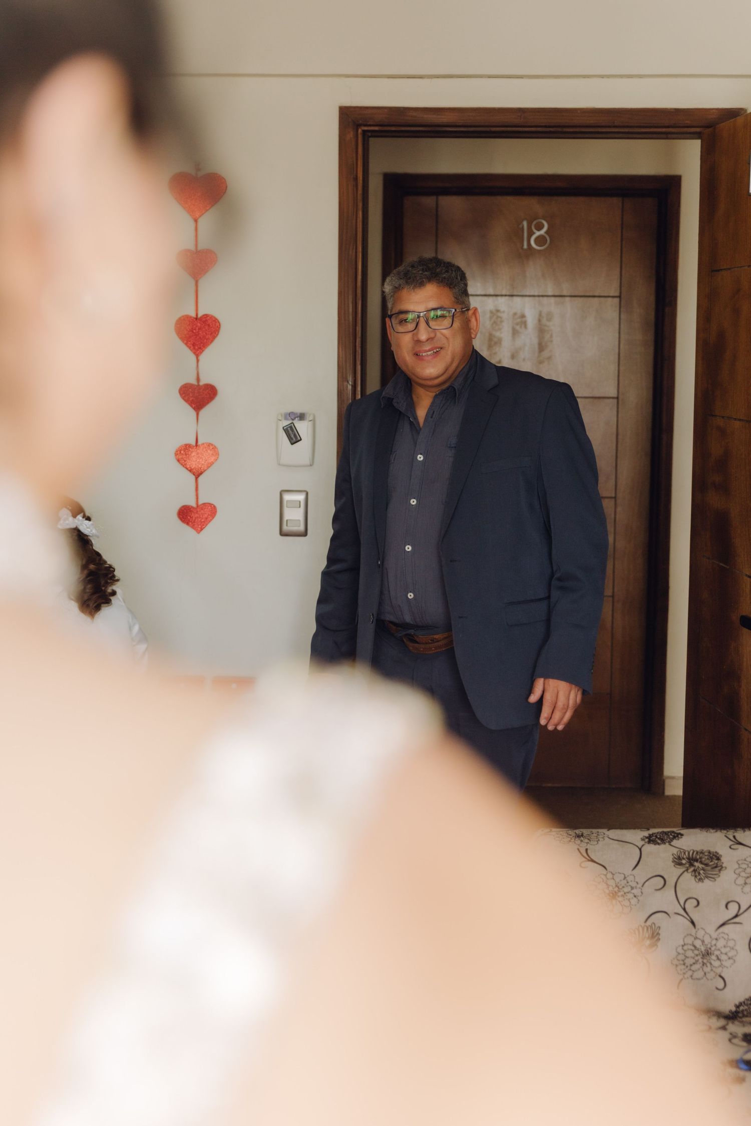 Father in navy blazer smiling at bride during emotional first look moment in decorated room.