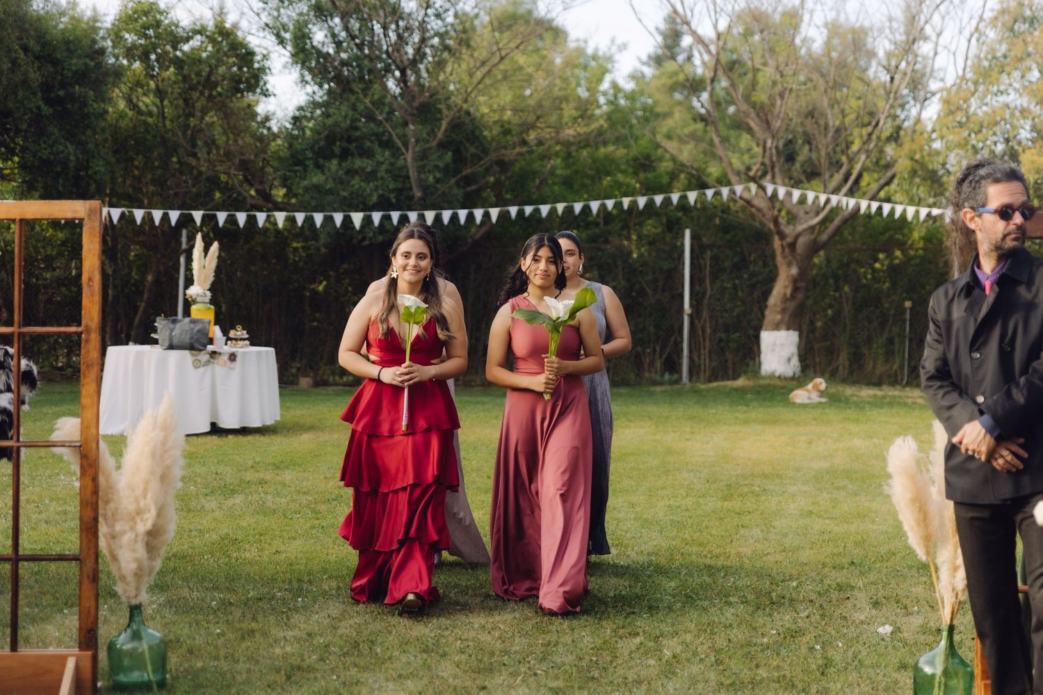 Three bridesmaids in red and mauve dresses carrying bouquets walking down an outdoor wedding aisle.