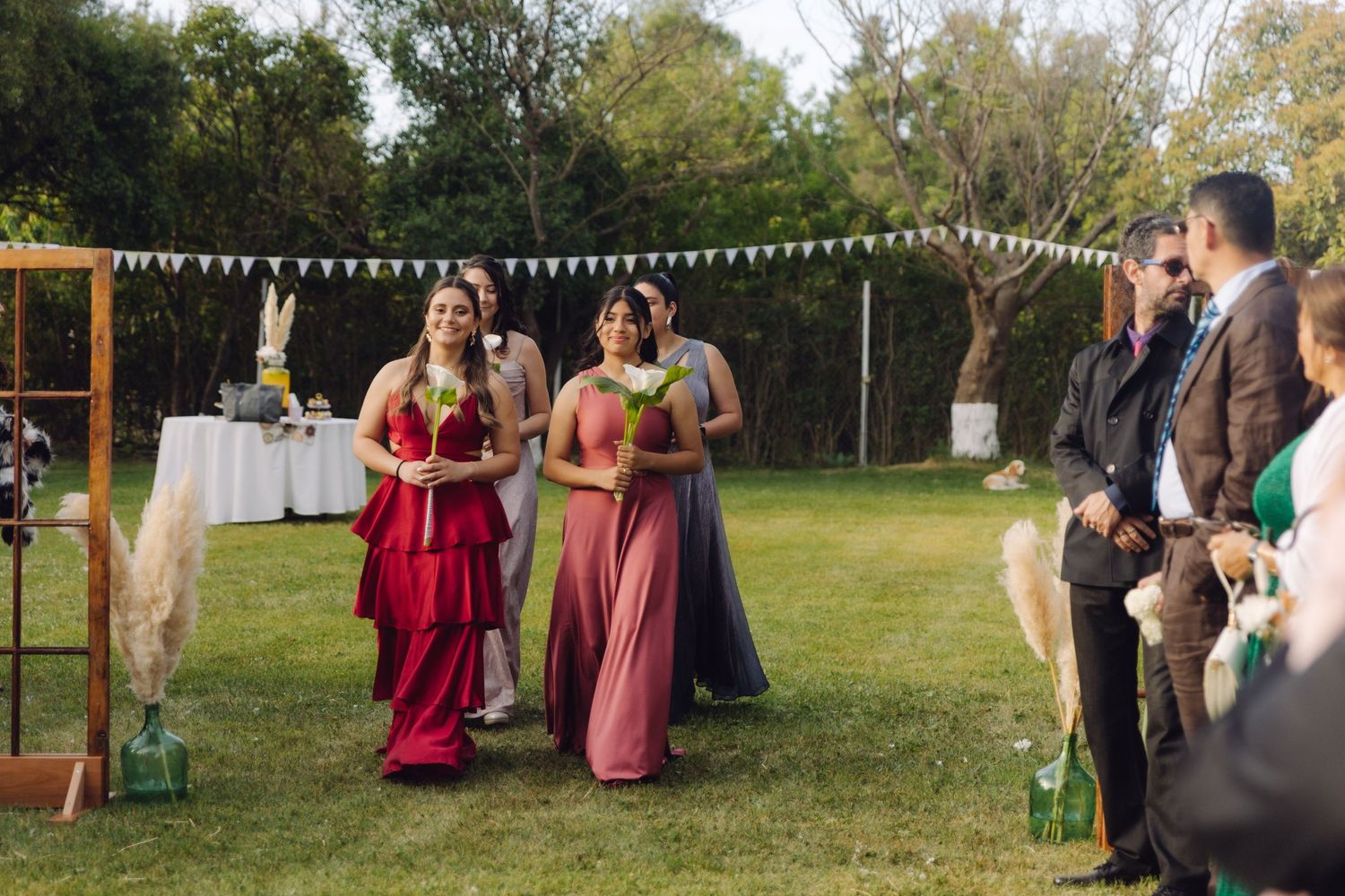 Bridesmaids in elegant red and dusty rose gowns processing down a garden wedding ceremony aisle.