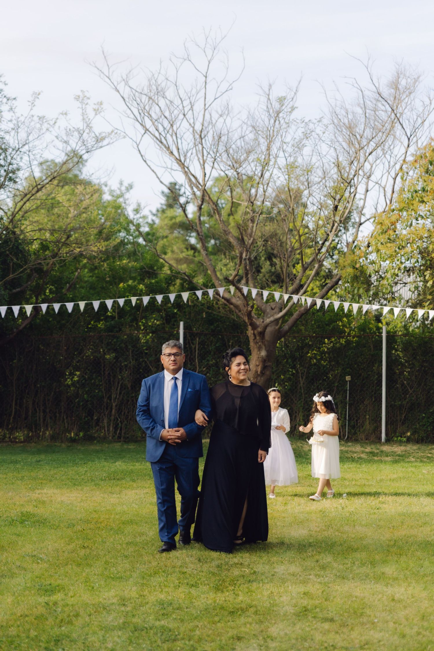 Couple in formal attire walking through a garden wedding venue with flower girl in background.