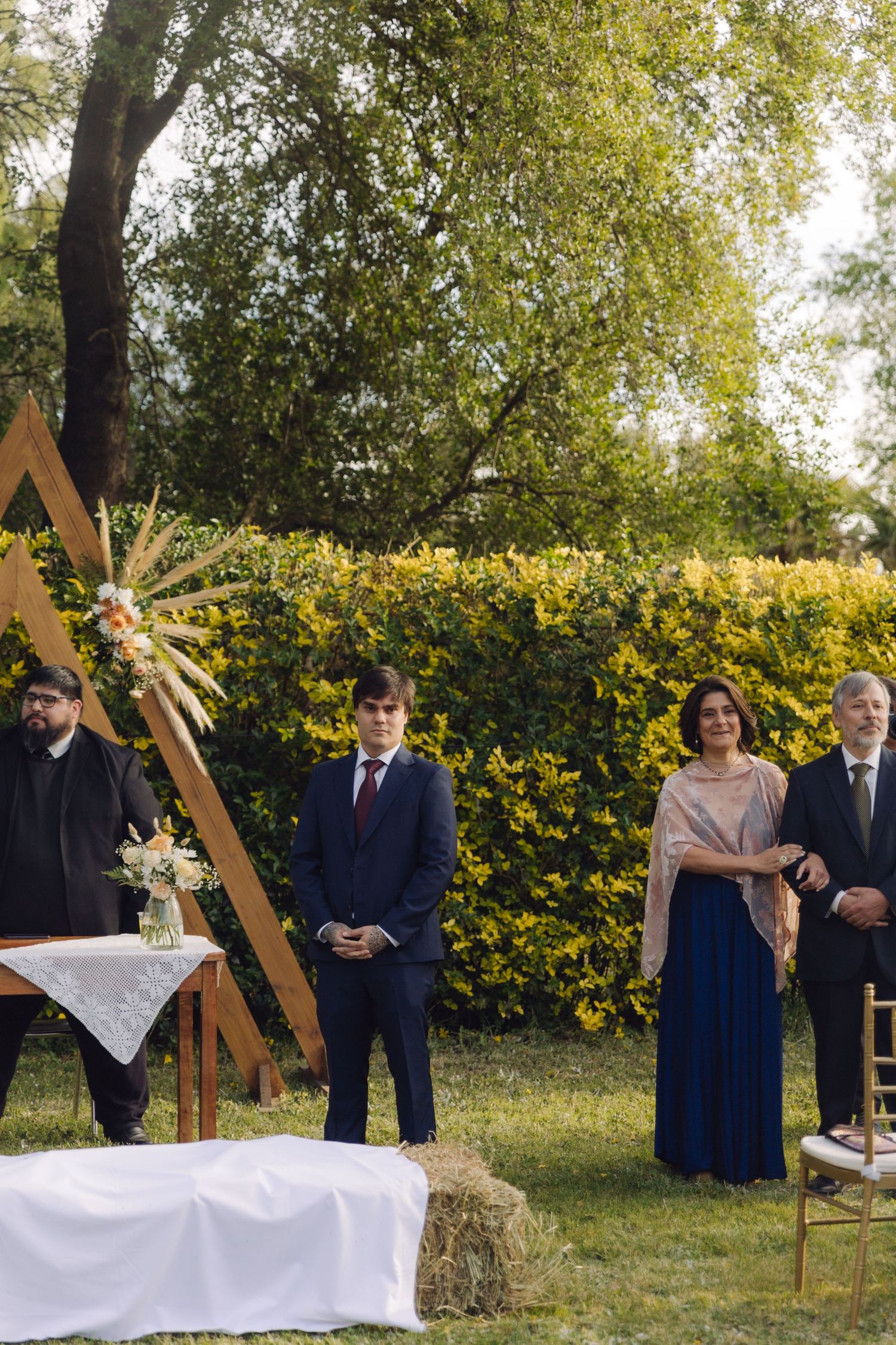 Groom in navy suit standing at outdoor altar with wooden triangular arch during wedding ceremony.