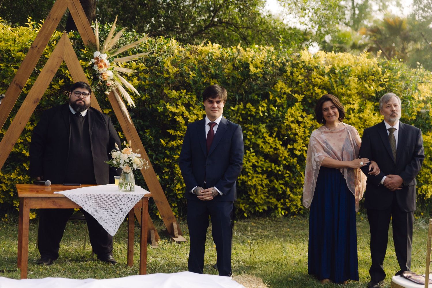 Outdoor wedding ceremony with officiant at wooden altar, groom in navy suit standing with two guests in a garden.