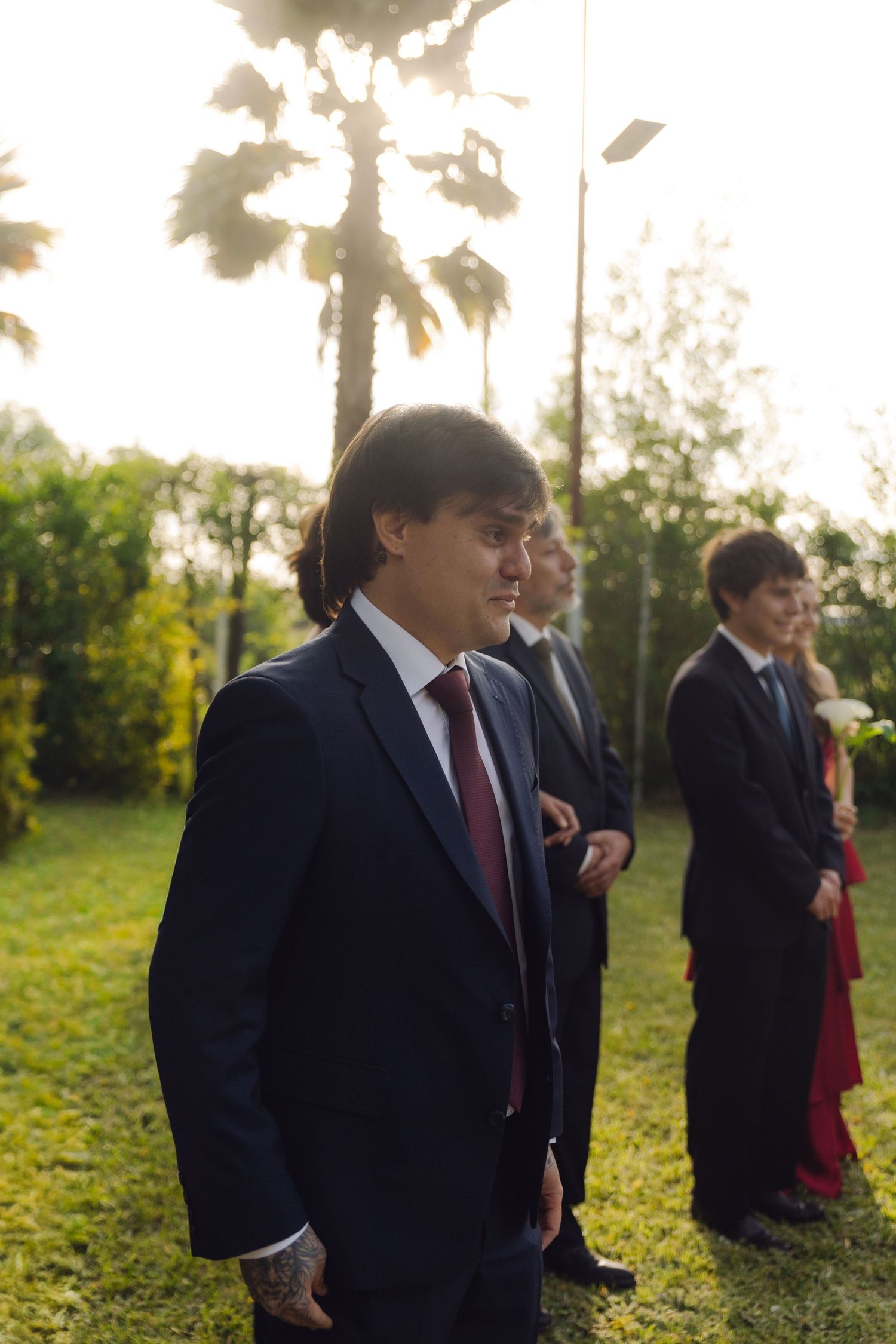 Groom in dark suit with burgundy tie standing outdoors at a garden wedding ceremony at sunset.