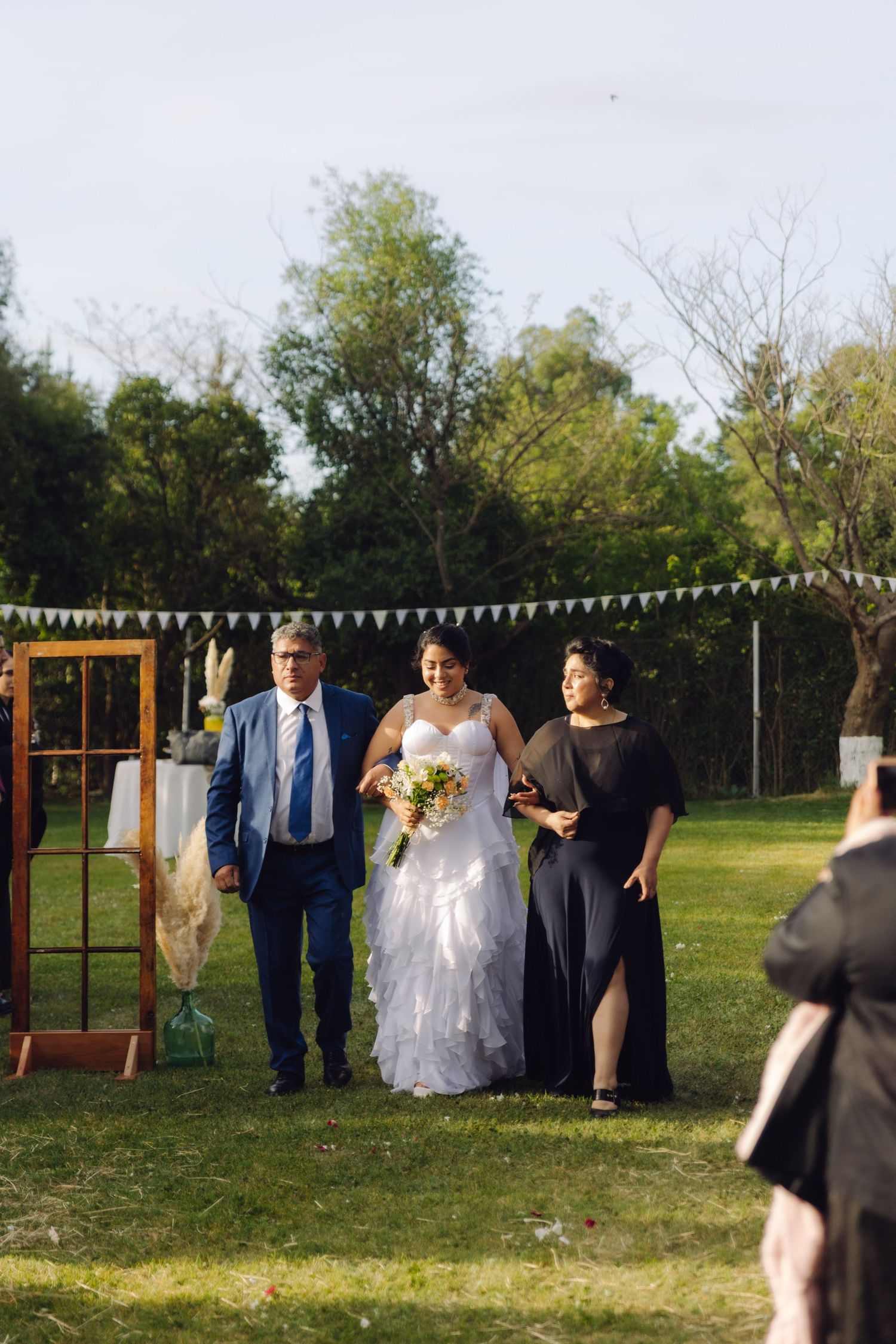 Bride in white ruffled gown walks down outdoor aisle escorted by father in blue suit and woman in black dress.