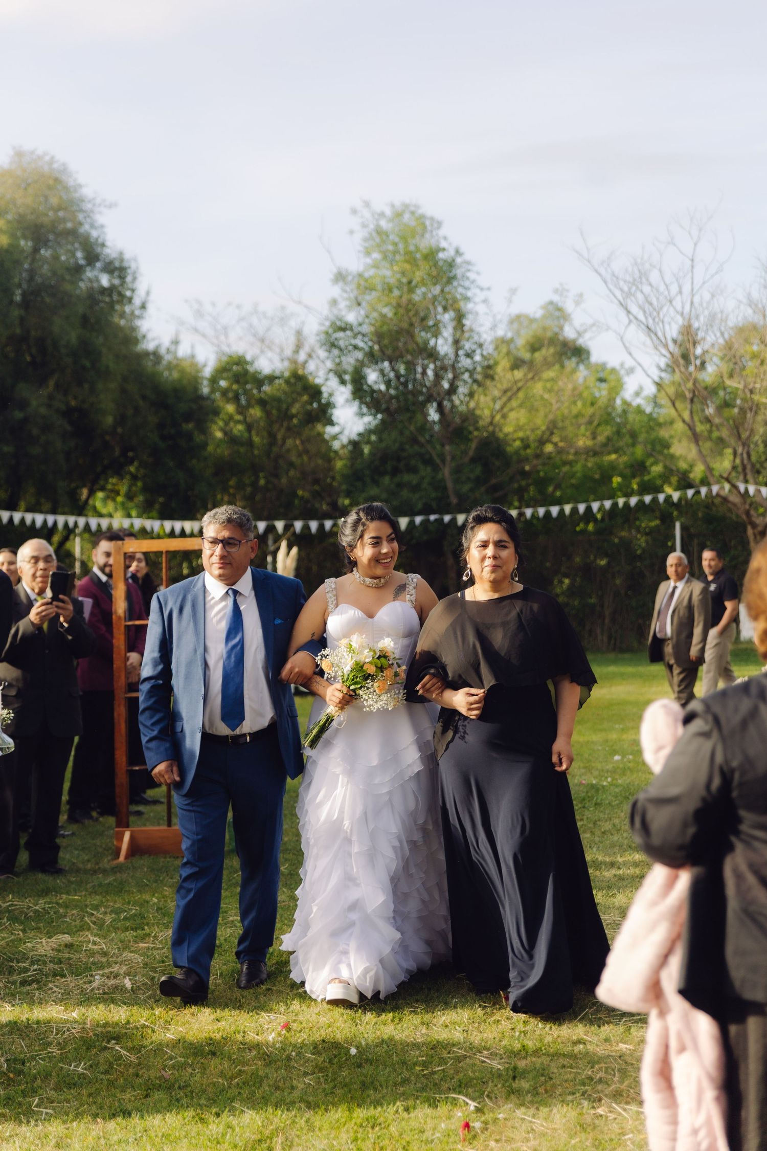 Bride holding colorful bouquet walks down grass aisle flanked by parents in blue suit and black dress outdoors.