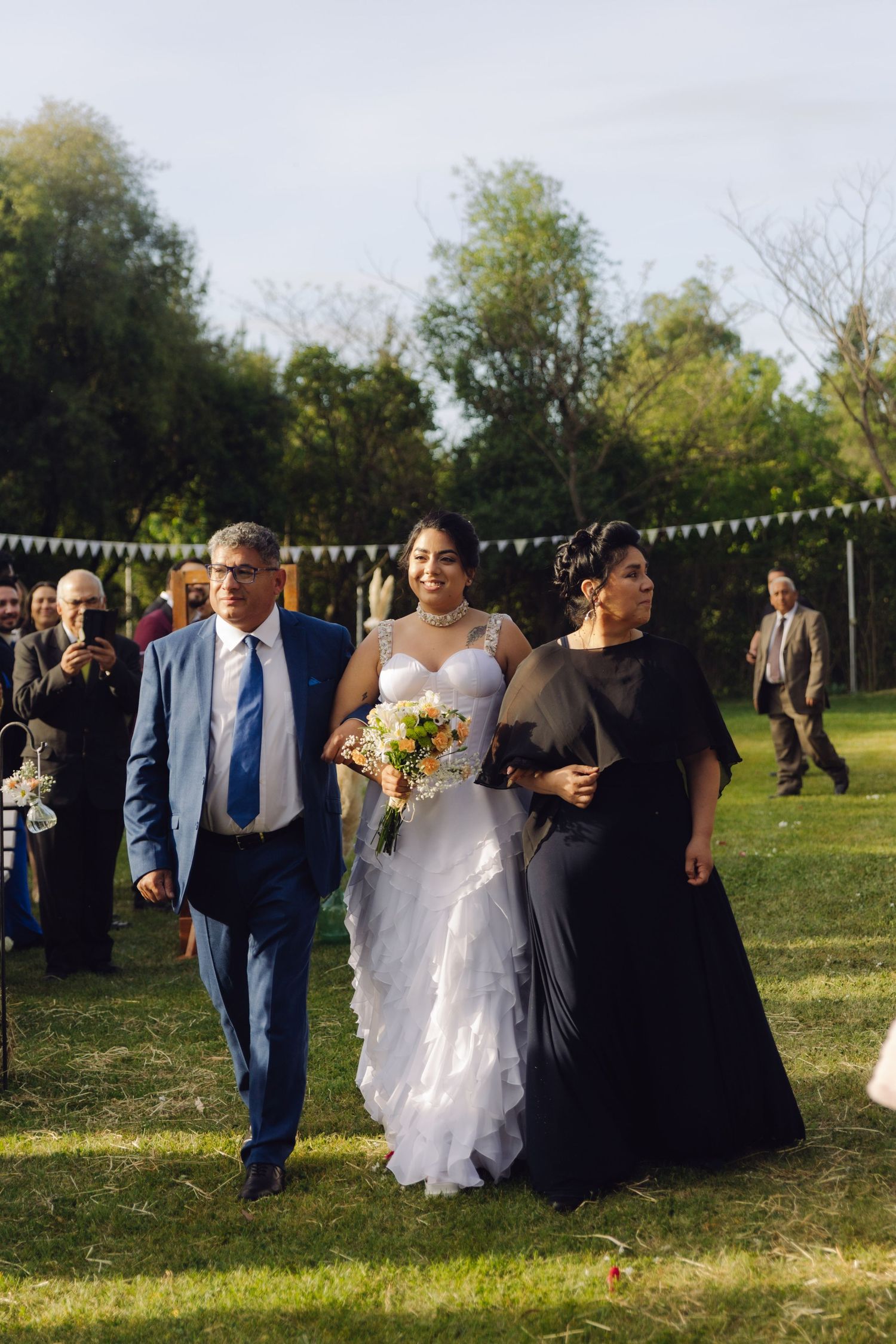 Bride in strapless white gown walks toward altar with parents through sunny outdoor garden wedding ceremony.