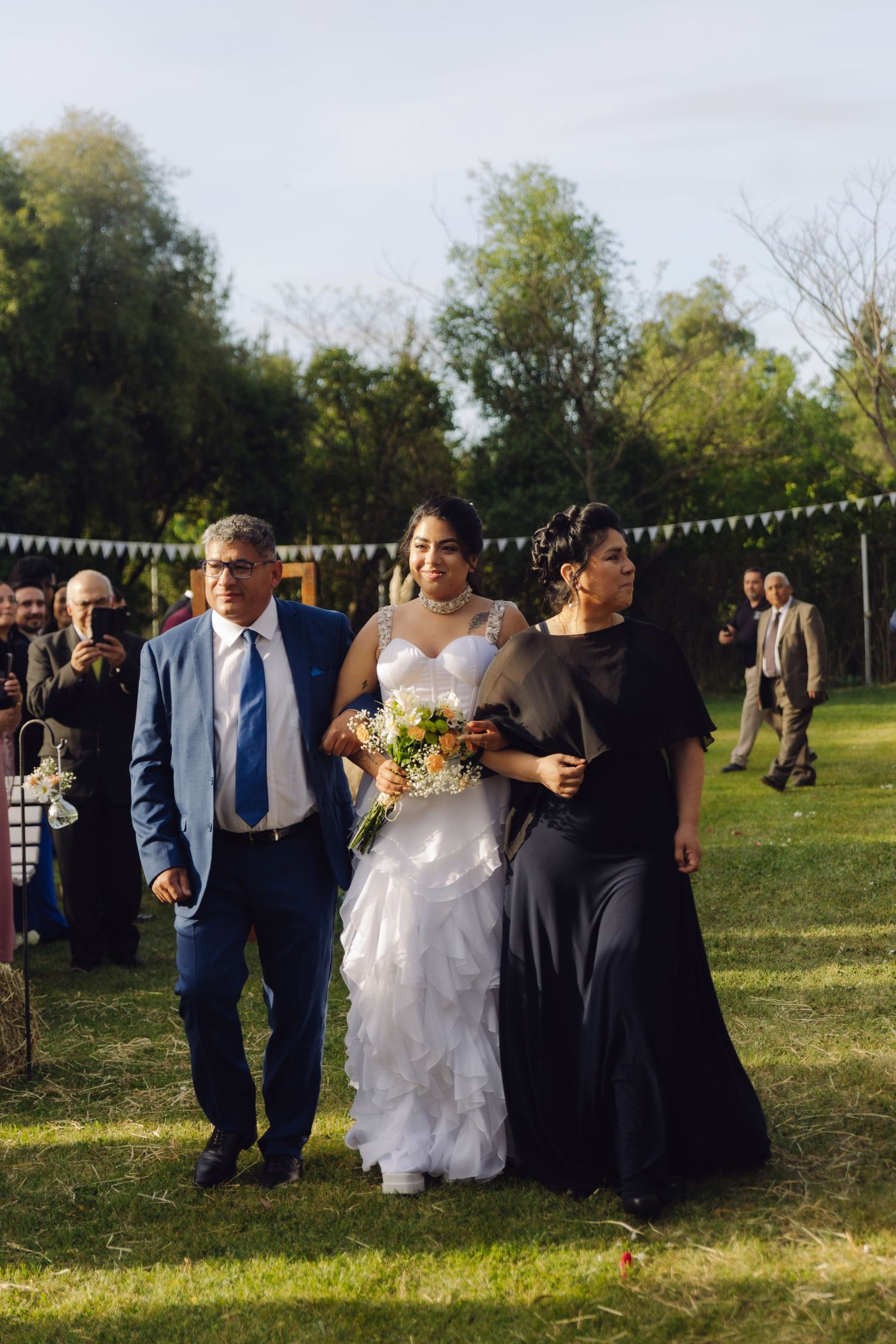 Close-up of bride with bouquet walking down aisle with father in blue suit and mother in black gown outdoors.
