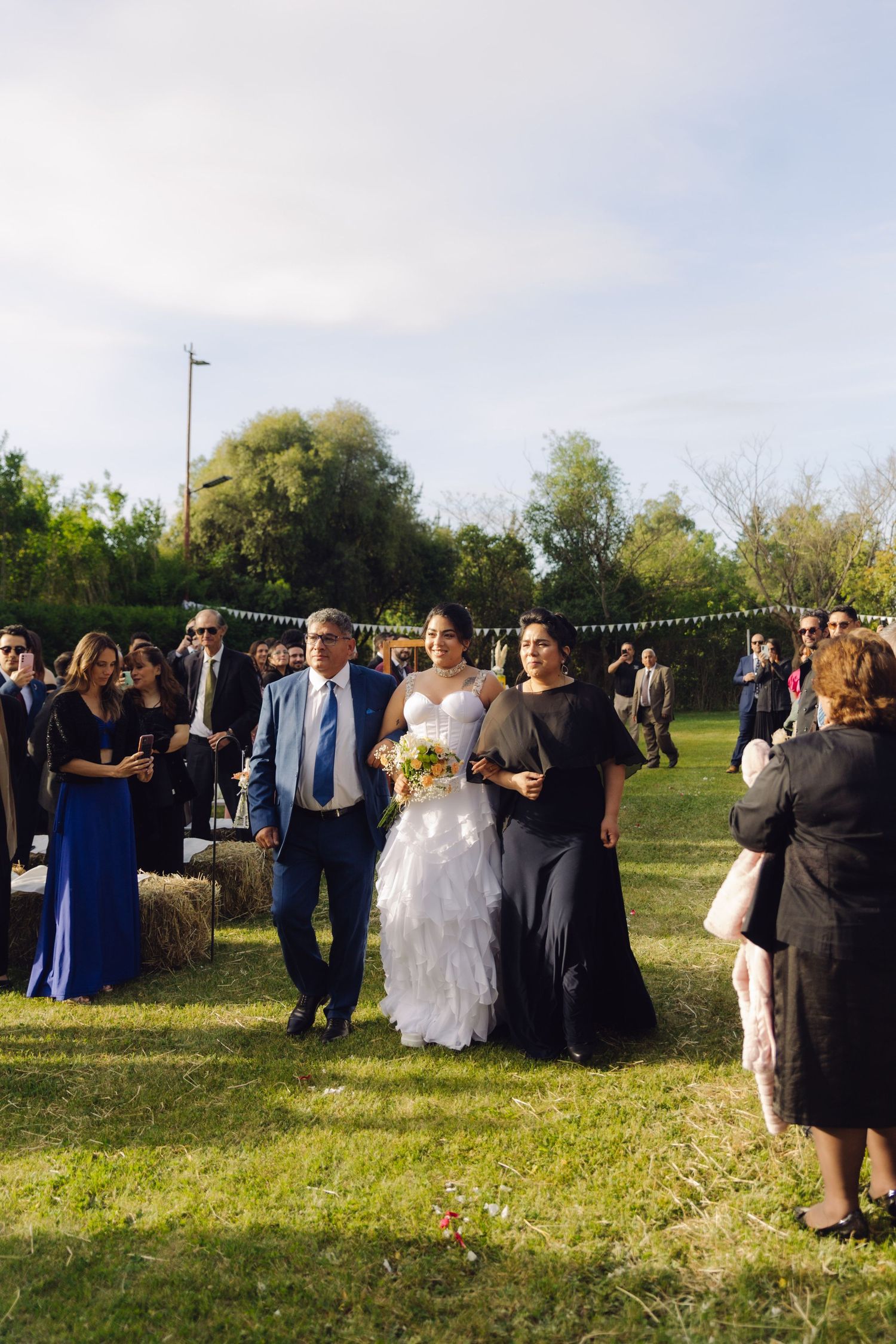 Bride and parents walk down outdoor grass aisle past seated and standing wedding guests on sunny day.