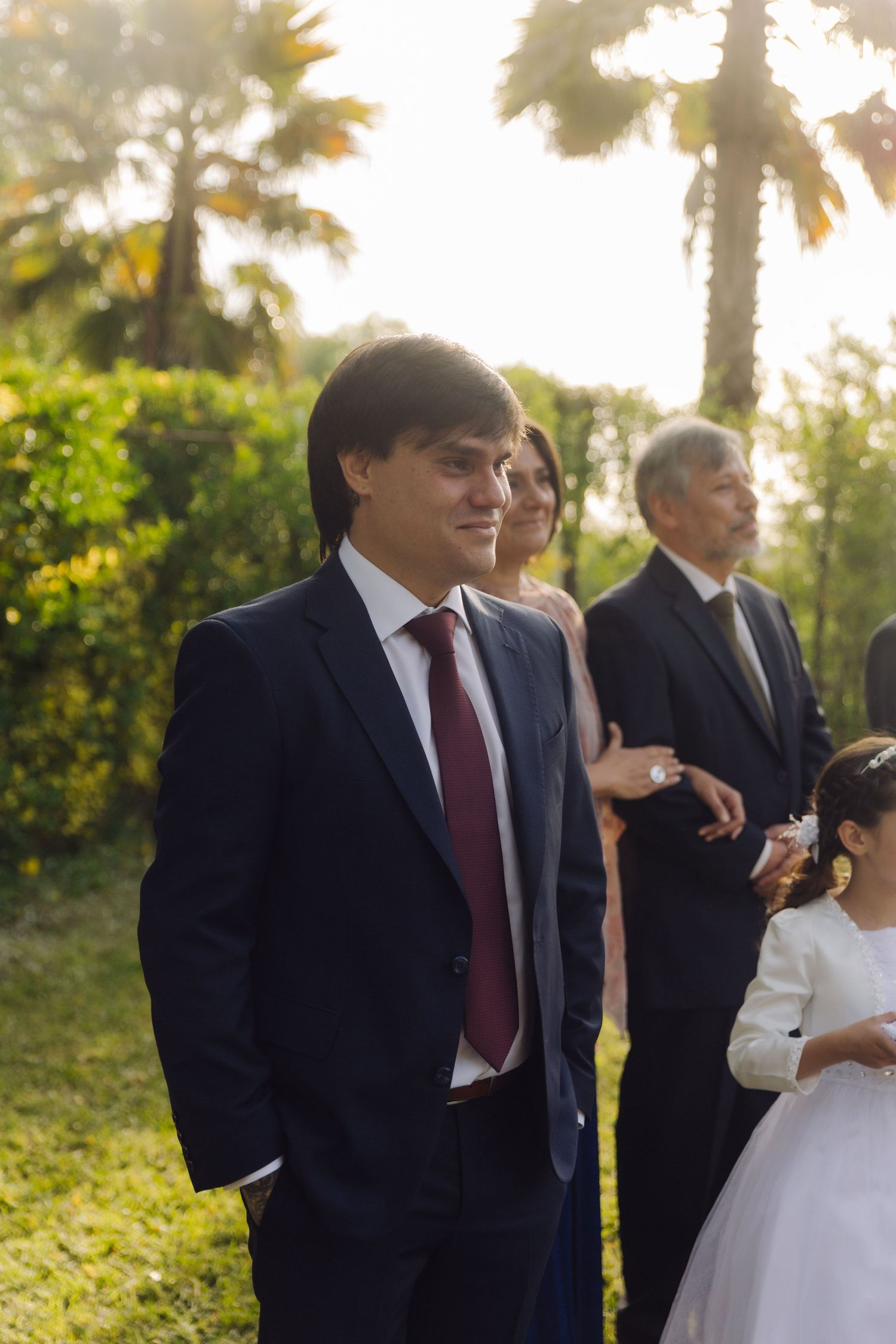 Groom in dark suit with burgundy tie stands outdoors at garden wedding ceremony with guests behind him.