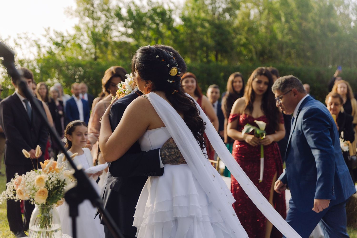 Bride and groom embrace at outdoor wedding altar surrounded by guests and bridesmaids in red dresses.